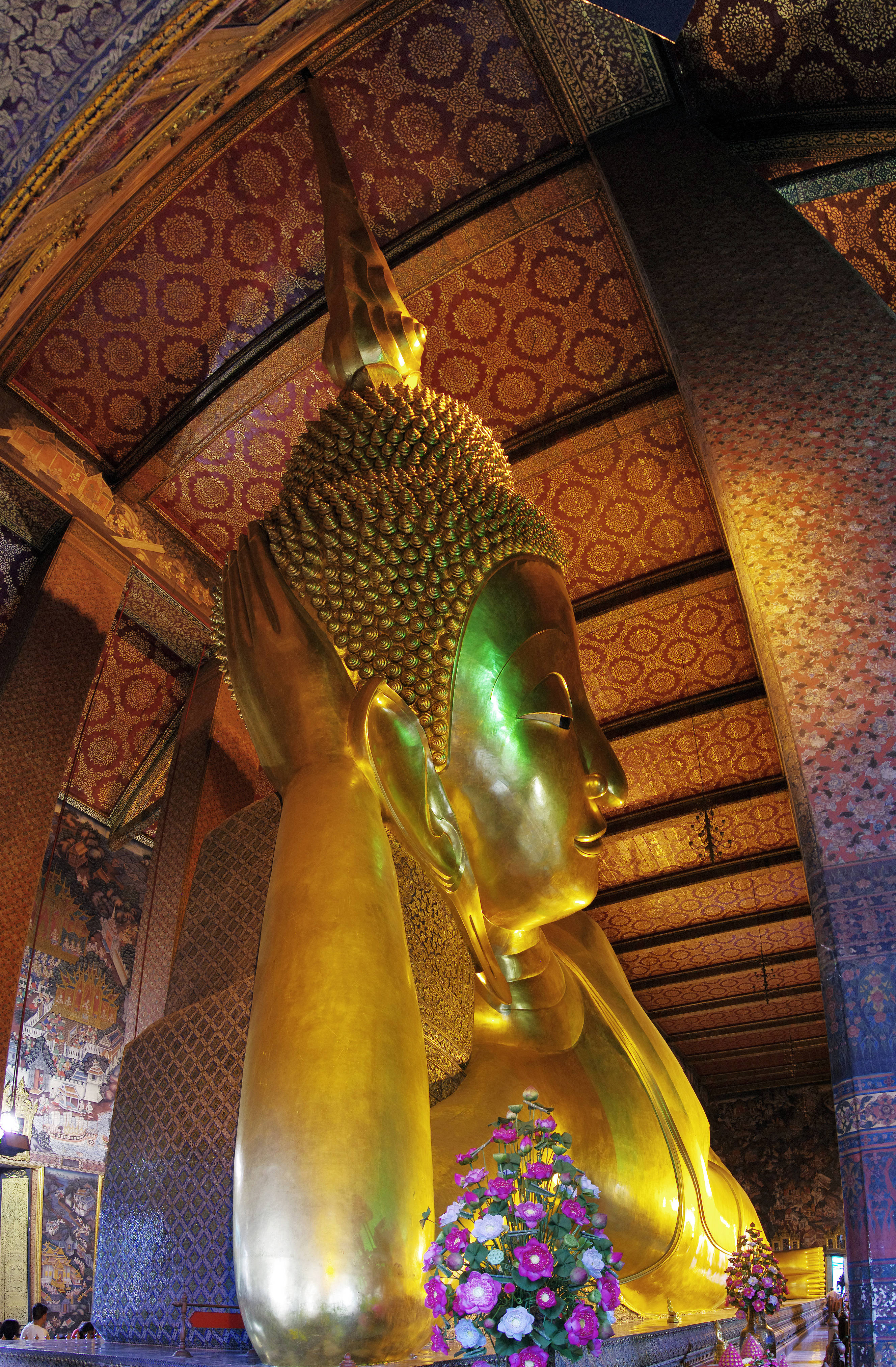Bangkok 桌布 壁紙 — 一尊巨大的金色臥佛在裝飾華麗的寺廟中安詳地躺著，被溫暖的光線照亮，底座上有色彩繽紛的花卉供品。
