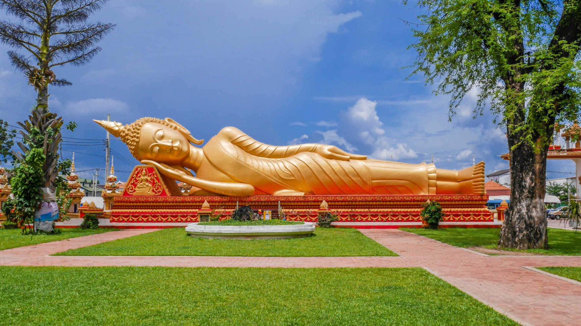 Laos 桌布 壁紙 — 一尊巨大的金色臥佛安詳地躺在紅色平台上，背景是晴朗的藍天和幾朵白雲。