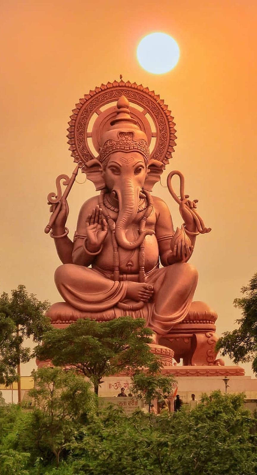 ganesh iPhone 桌布 壁紙 — 一尊巨大的金色象神雕像在燦爛的陽光下靜靜地坐著，周圍綠樹成蔭，環繞著底座。