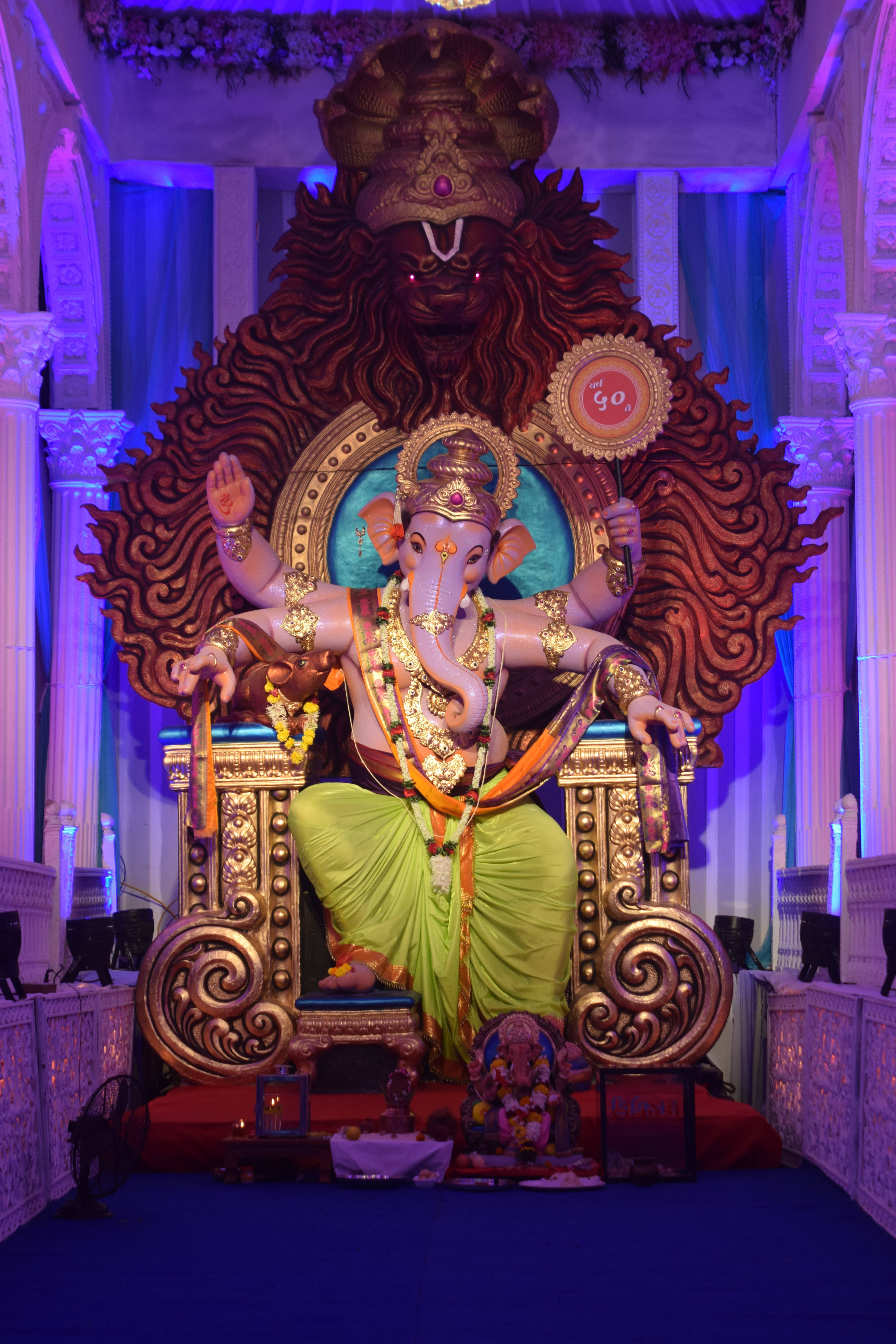 hindu iPhone 桌布 壁紙 — 一尊巨大的金色象頭神像坐在華麗的寶座上，背景有獅子頭，在紫色舞台燈光下。