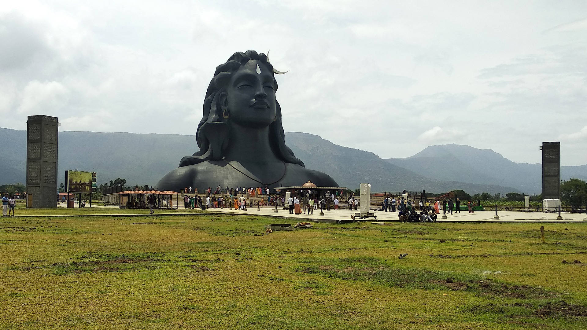 shiv shankar 桌布 壁紙 — 一尊巨大的黑色石雕濕婆神像，帶有新月，點綴著一片草地，遠處有山脈，天空多雲。