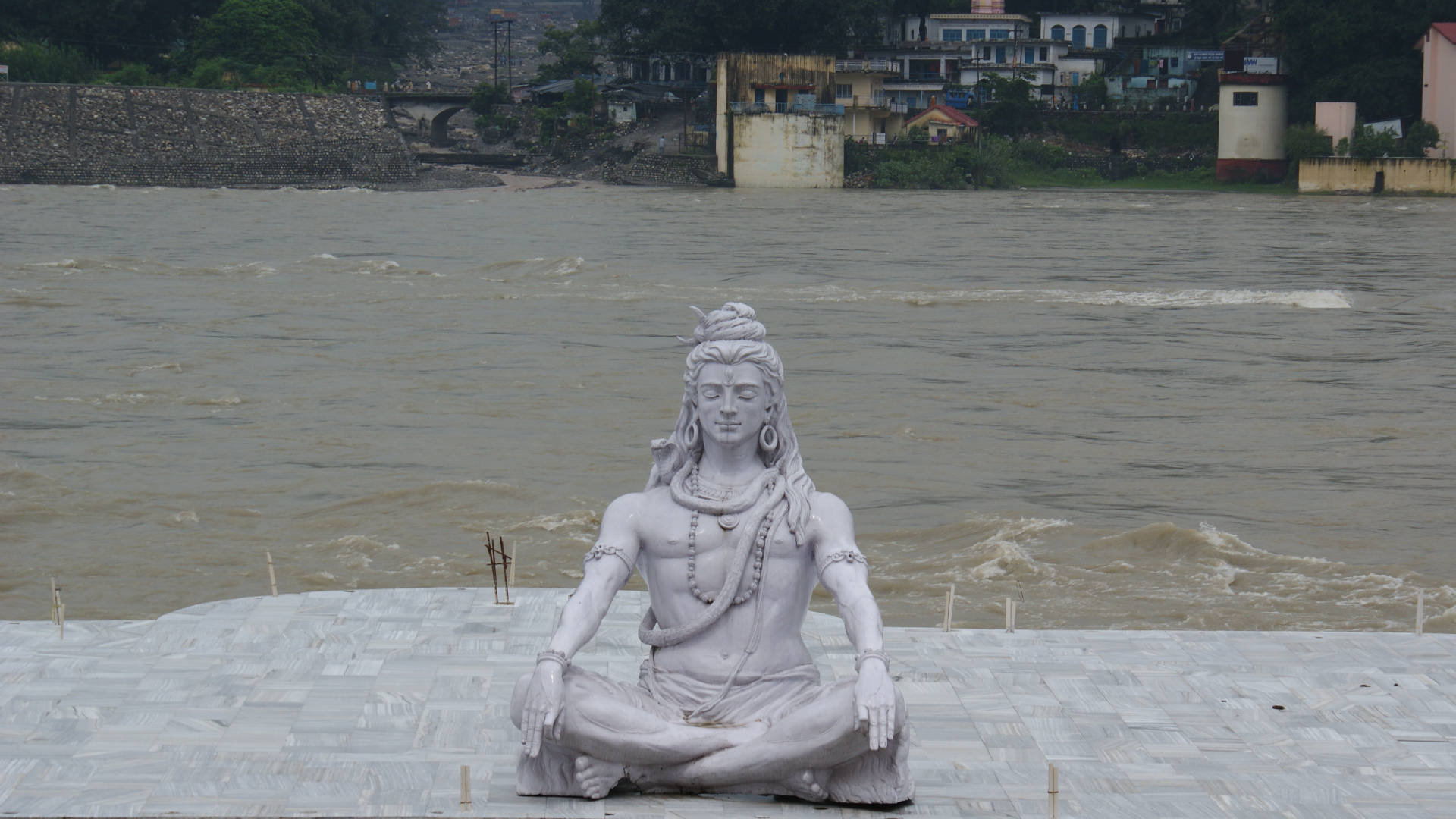 Hindu God 桌布 壁紙 — 一尊白色石雕的冥想神祇，盤腿坐在河邊的瓷磚平台上，渾濁的棕色河水奔流而過。
