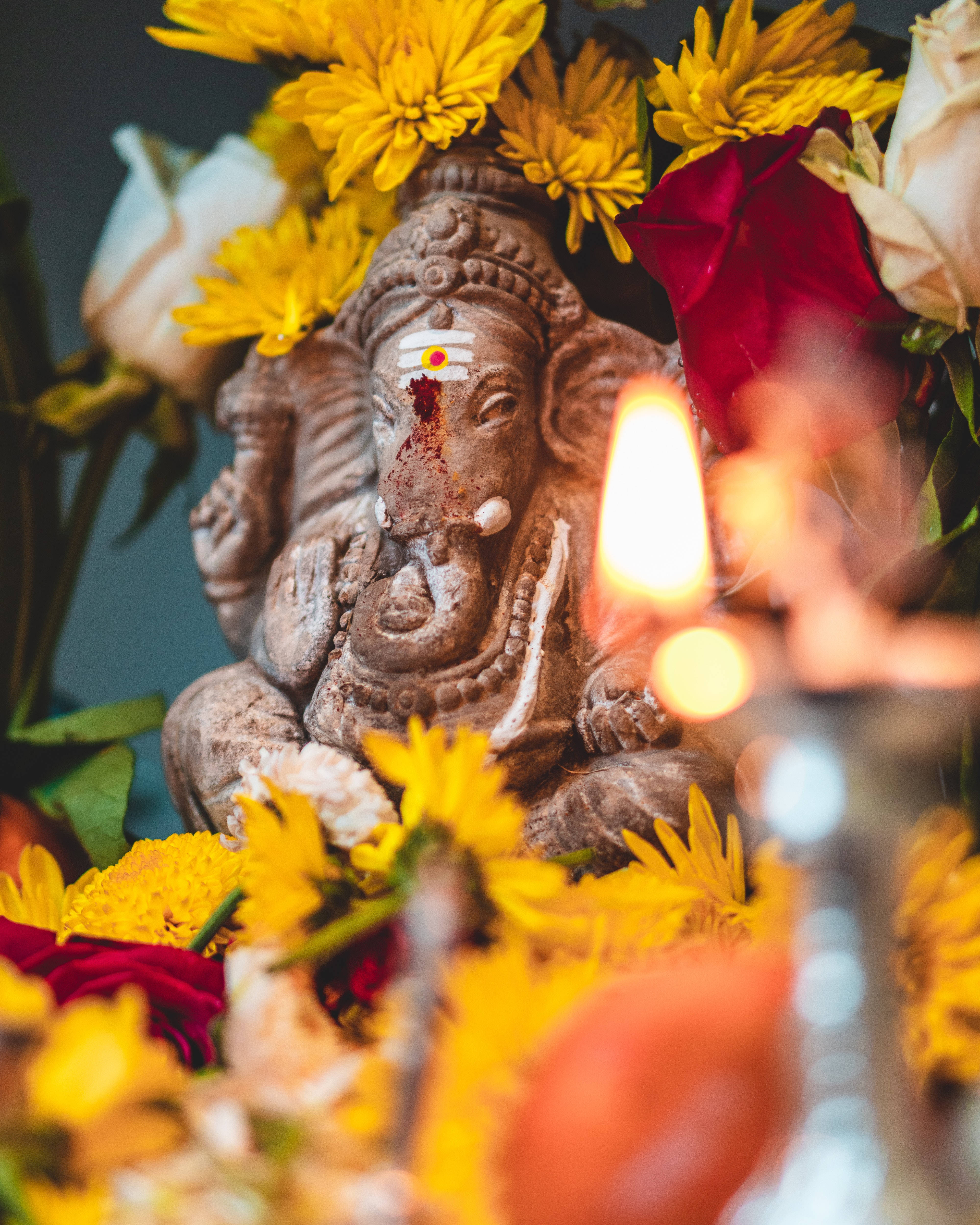 Ganesh 桌布 壁紙 — 一尊石雕象神像，周圍環繞著鮮豔的黃色和紅色花朵，前景中有一根發光的蠟燭，焦點柔和。