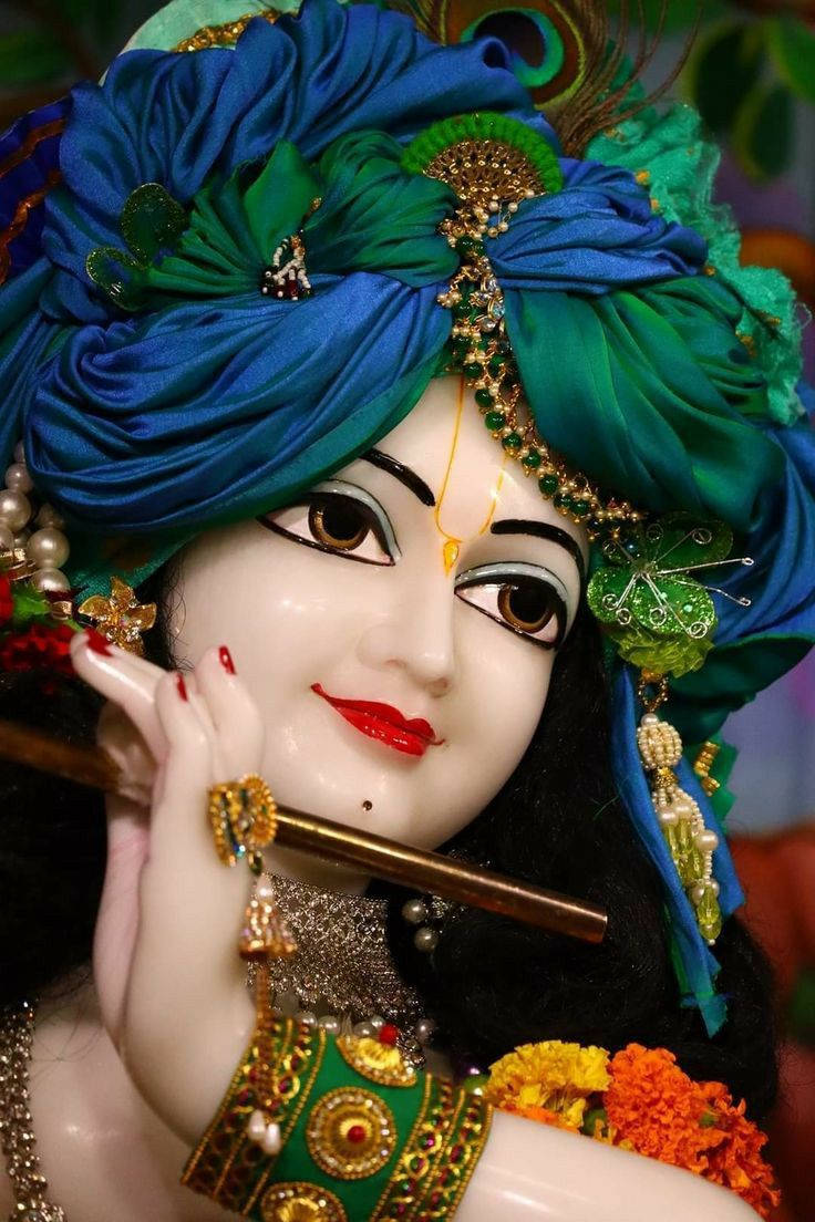 Hindu God 桌布 壁紙 — 一尊神聖的克里希納雕像，頭戴鮮豔的藍綠色頭飾，帶著寧靜的微笑吹奏長笛。