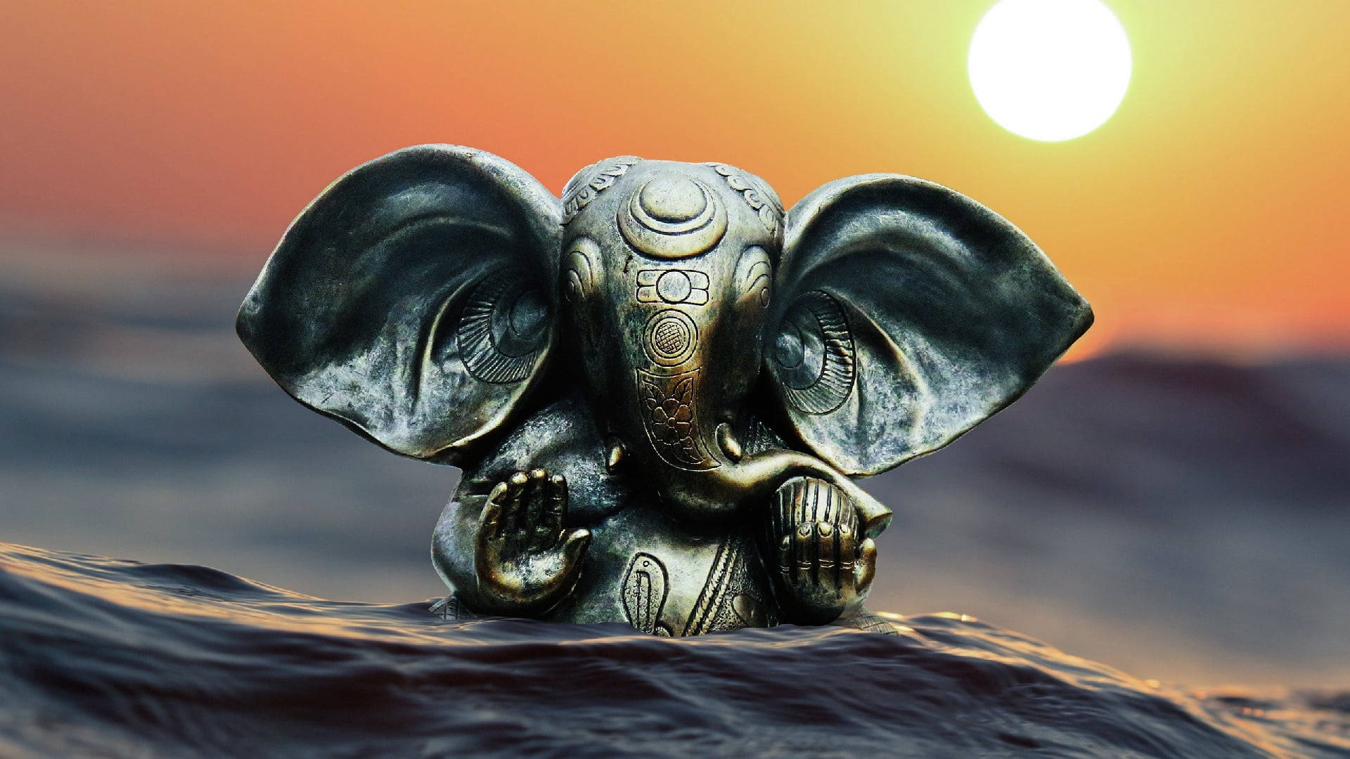 Hindu God 桌布 壁紙 — 一尊精細的青銅象神雕像在夕陽下從海浪中浮現，背後有發光的太陽。