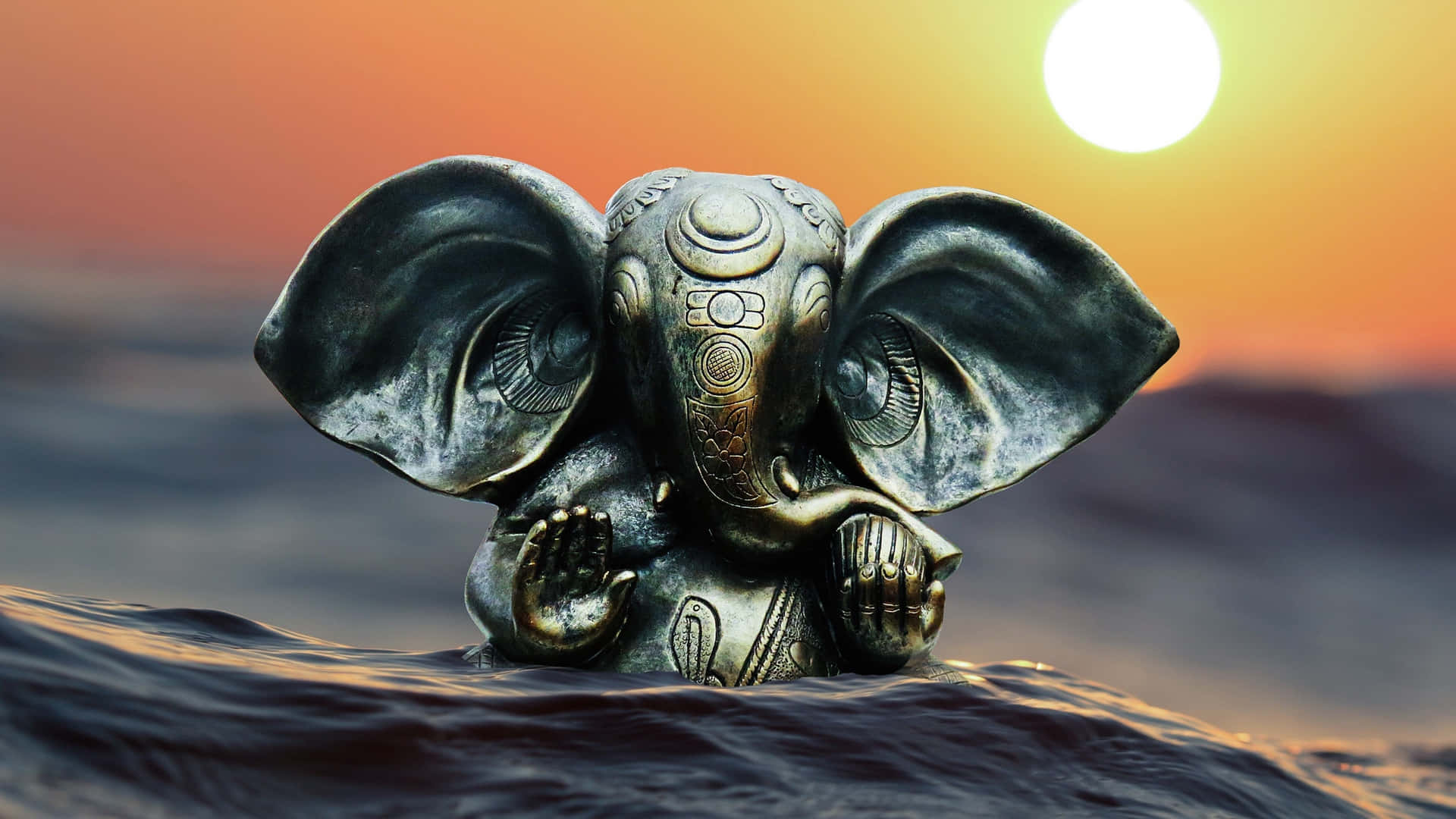 Ganesh 桌布 壁紙 — 一尊精細的青銅象神雕像從暗水中浮現，背後是鮮豔的橙色夕陽。