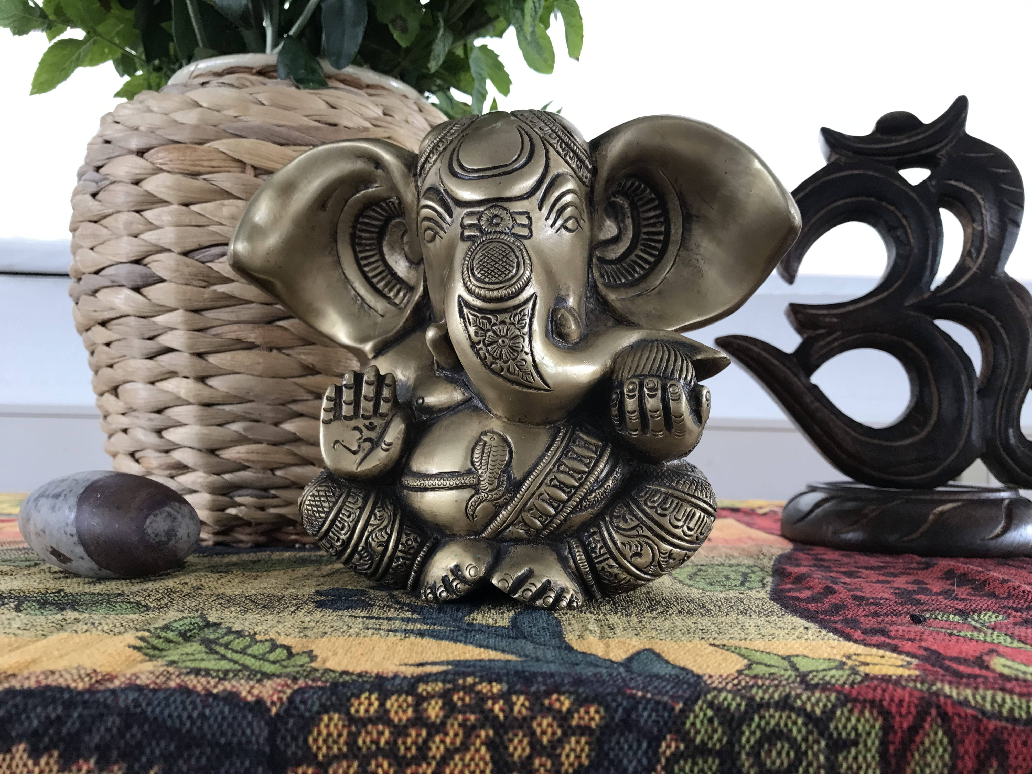 cute ganesha 桌布 壁紙 — 一尊精細的黃銅象神雕像放在色彩繽紛的圖案布上，旁邊有一個編織籃子和木製的Om符號。