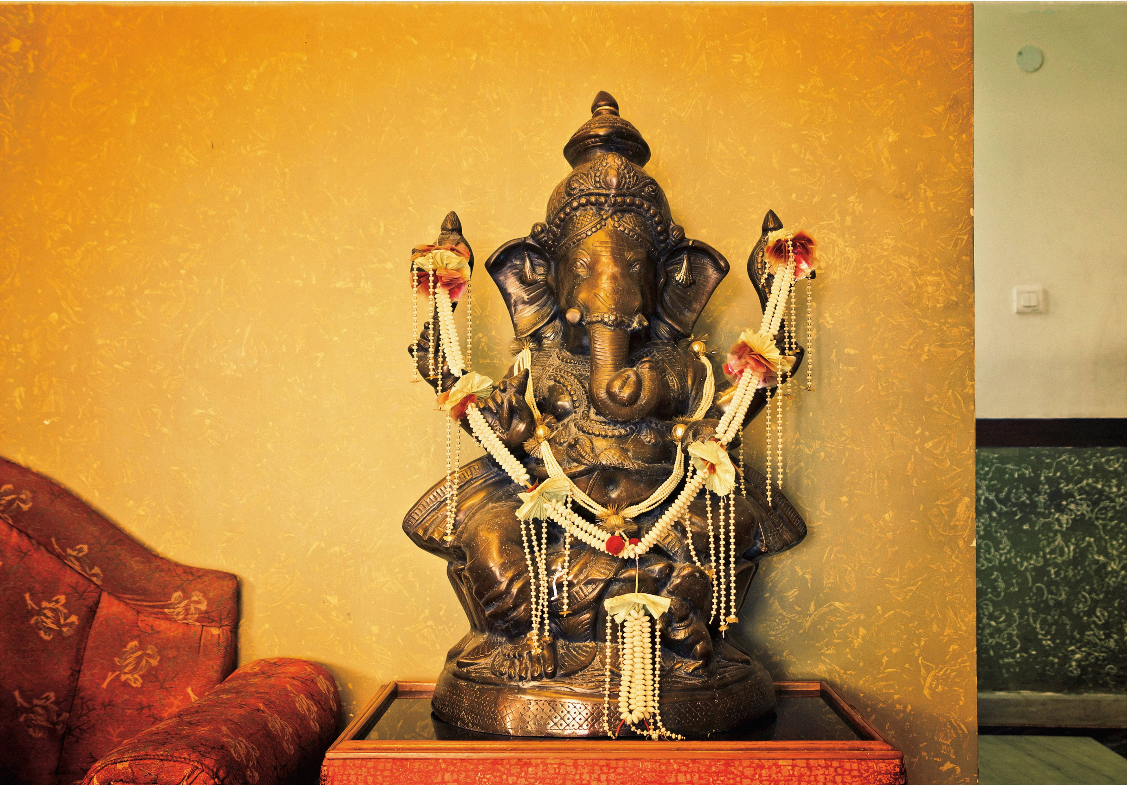 Ganesh 桌布 壁紙 — 一尊精緻的青銅象神雕像，飾以白色和紅色花環，放置在木桌上，背景是帶有紋理的金色牆壁。
