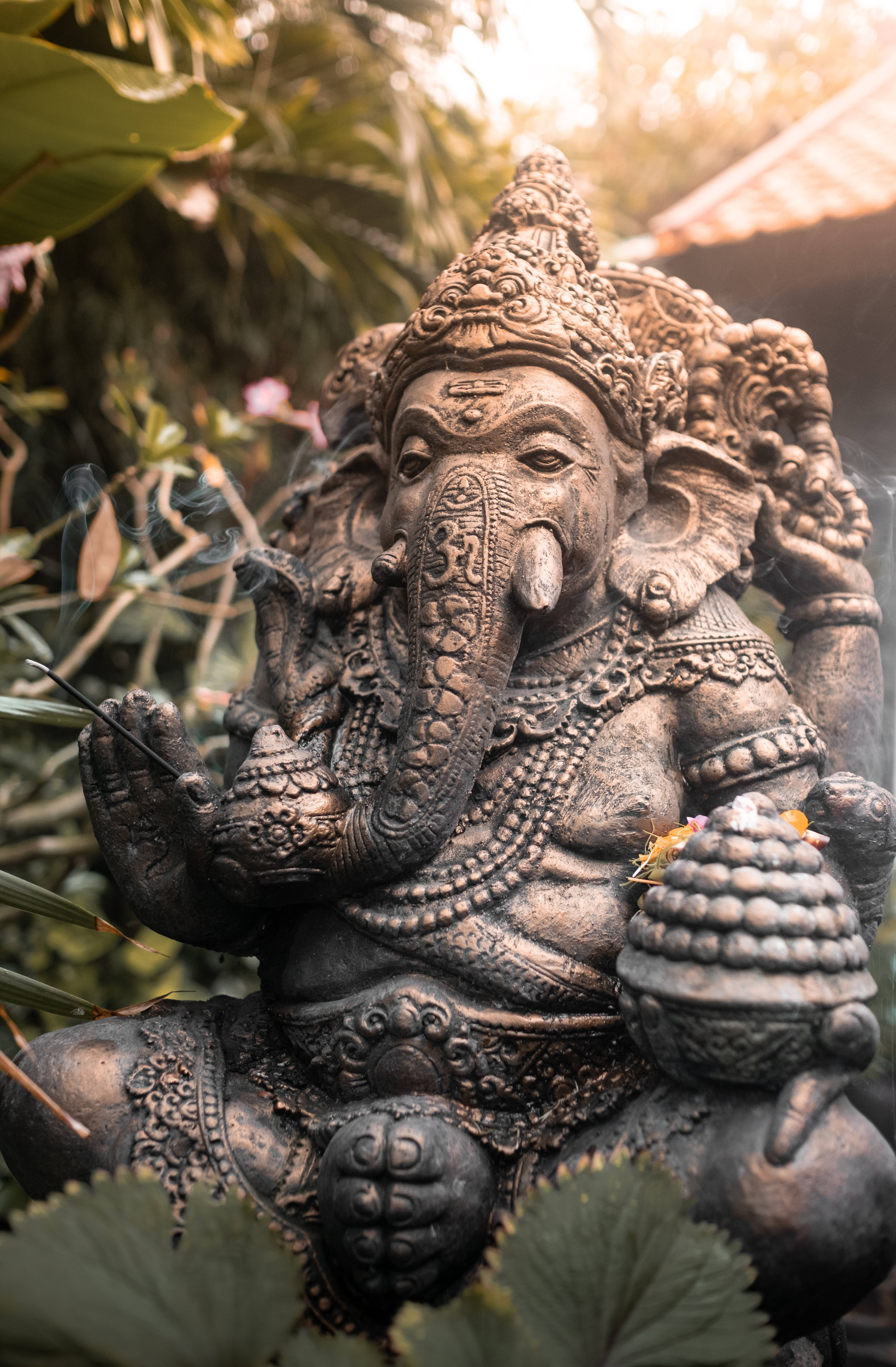 ganesh iPhone 桌布 壁紙 — 一尊精緻的青銅象神雕像安詳地坐在茂密的綠葉中，柔和的金色陽光穿過樹葉灑落。