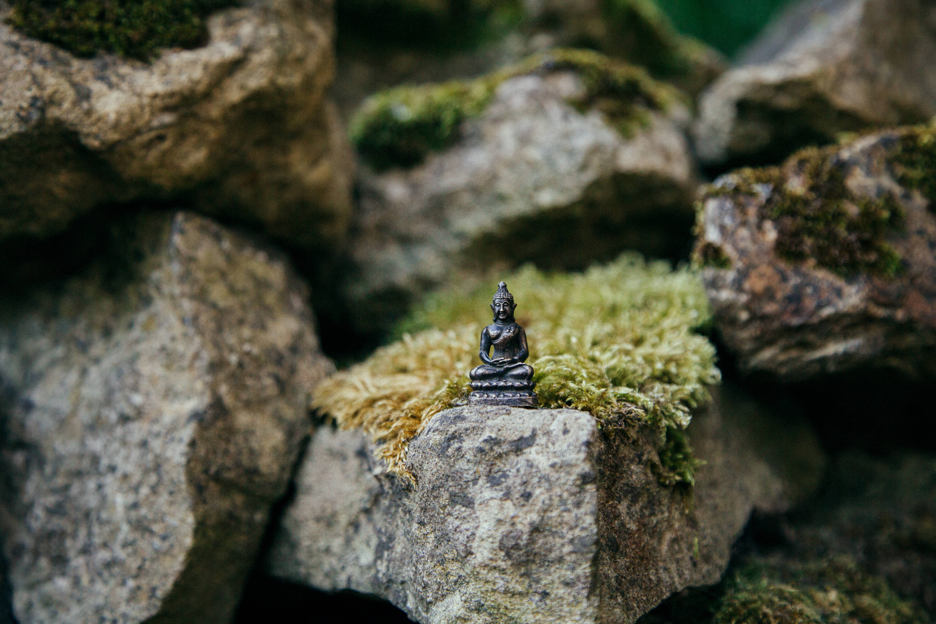 佛陀 桌布 壁紙 — 一尊細小的青銅佛像安詳地坐在長滿青苔的石頭上，周圍是粗糙的石塊。