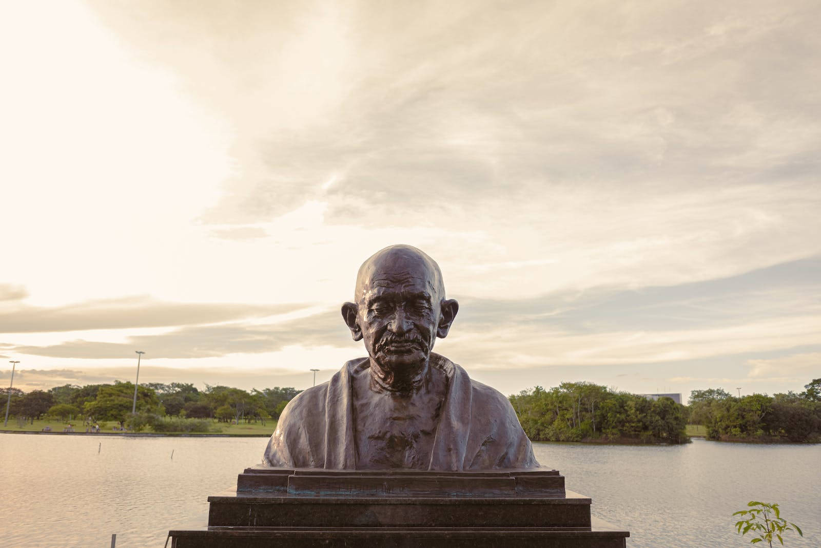 Mahatma Gandhi 桌布 壁紙 — 一尊聖雄甘地的銅製半身像莊嚴地矗立在寧靜湖畔的基座上，背景是柔和的金色日落天空。