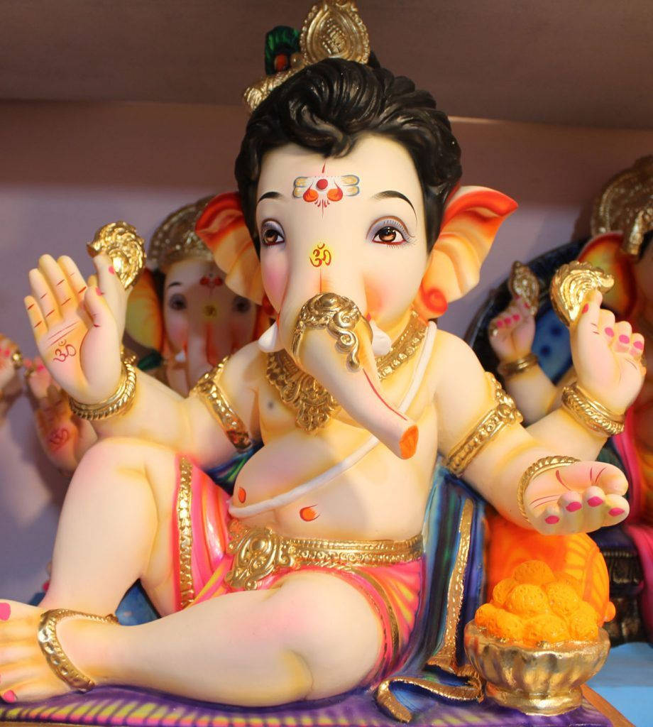cute ganesha 桌布 壁紙 — 一尊色彩繽紛的塑膠象神像，飾有金色珠寶和粉紅色服飾，在其他偶像中顯眼地坐著。