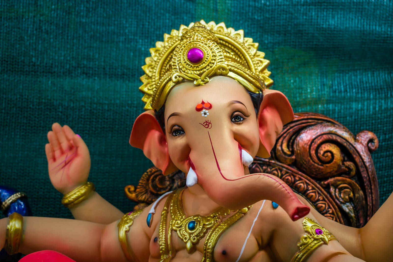 Vinayaka 桌布 壁紙 — 一尊色彩繽紛的象神像，飾有黃金珠寶和紫色寶石皇冠，背景為帶紋理的青綠色。