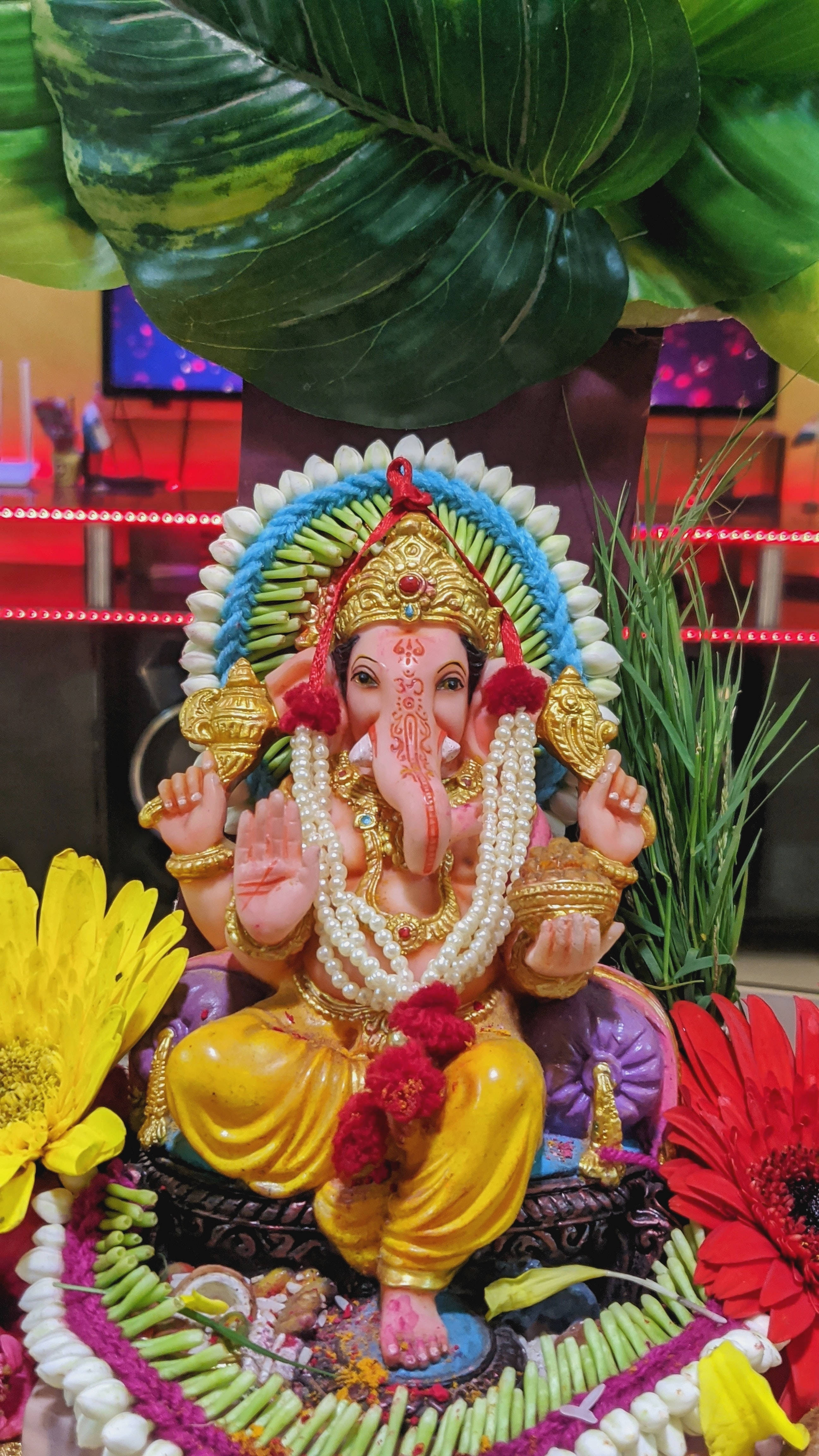 hindu iPhone 桌布 壁紙 — 一尊色彩繽紛的象神像，飾有黃金首飾和鮮花，坐在一片大綠葉下，周圍環繞著鮮豔的花朵。