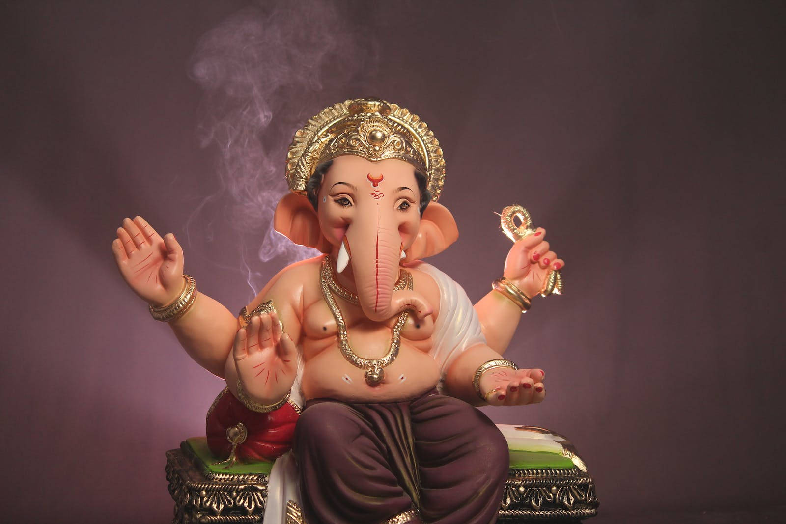 Vinayaka 桌布 壁紙 — 一尊色彩繽紛的象神像威嚴地坐在華麗的寶座上，背後有煙霧升起，襯托著深紫色的背景。