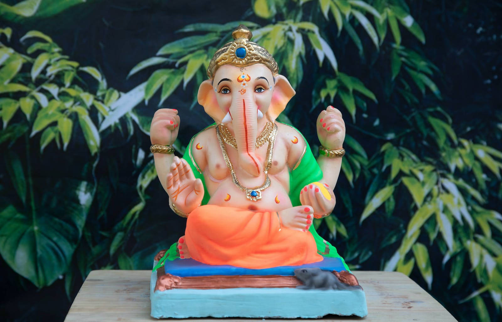 Vinayaka 桌布 壁紙 — 一尊色彩繽紛的象神像安詳地坐在基座上，背景是茂密的綠色樹葉。