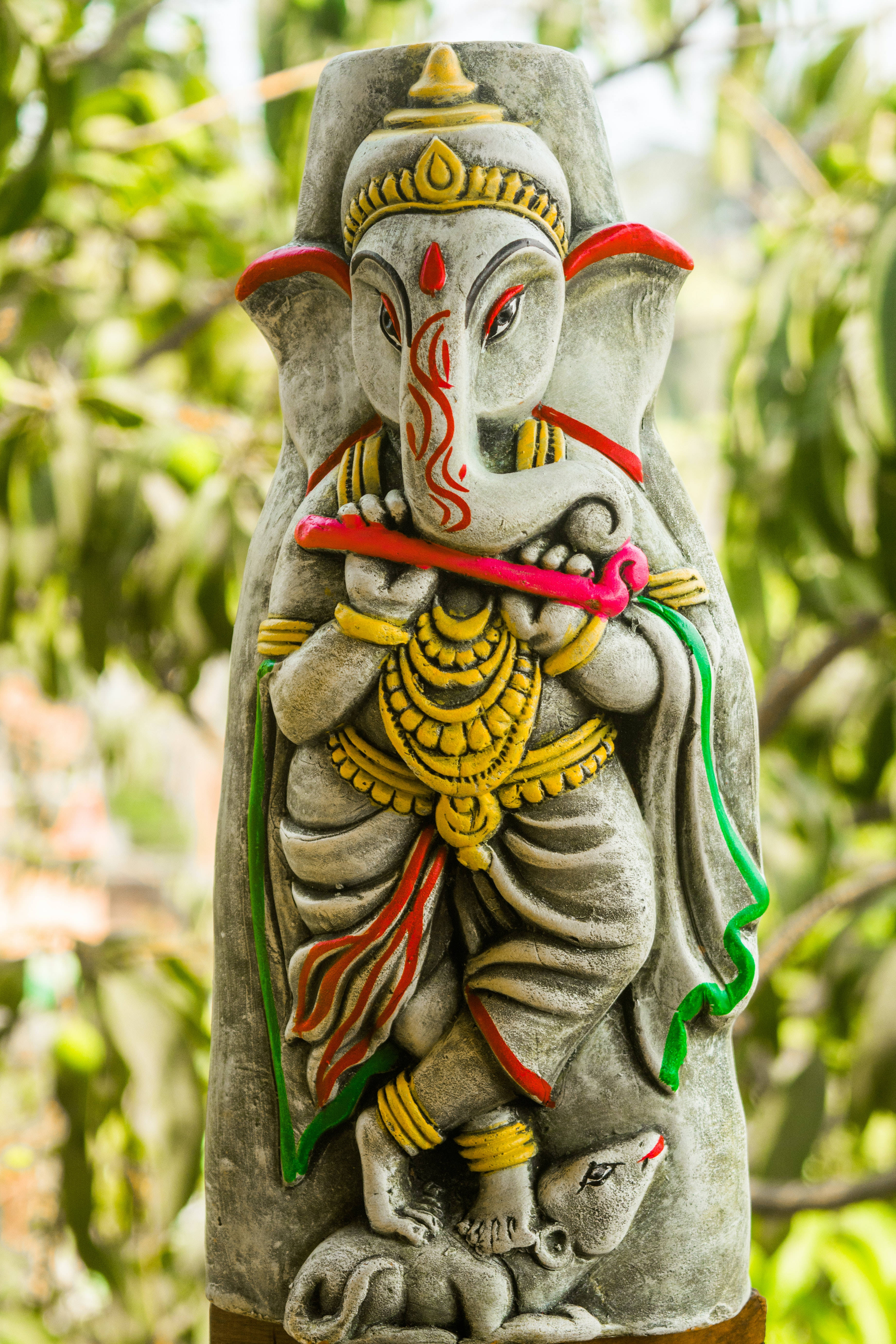 ganesh 4K 桌布 壁紙 — 一尊色彩繽紛的象頭神石像，紅眼睛，戴金飾，矗立在茂密的綠葉叢中。