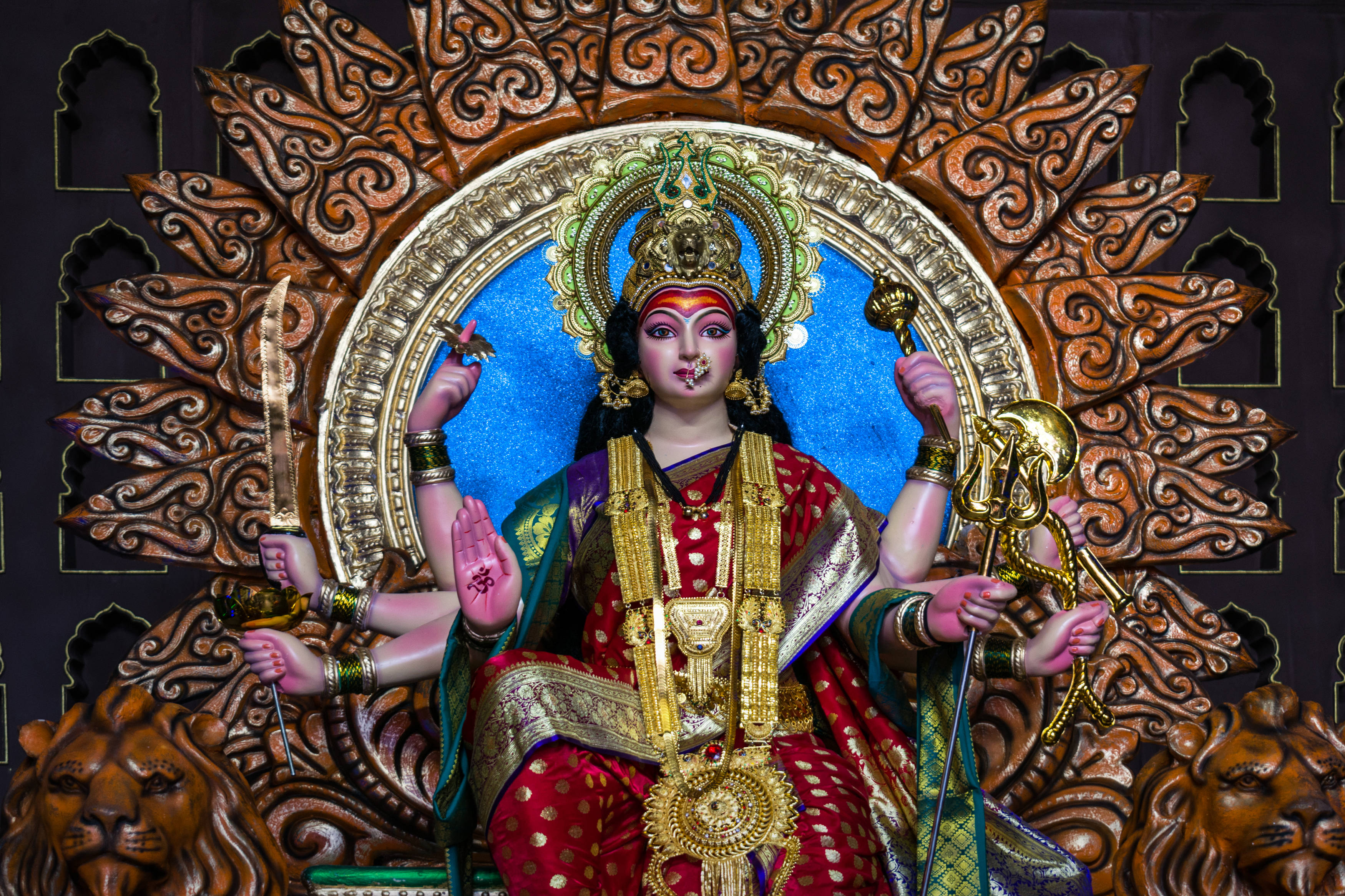 Navratri 桌布 壁紙 — 一尊色彩鮮明、細節豐富的印度教女神杜爾迦雕像，裝飾著紅色和金色，坐在有精緻木雕的獅子寶座上，手持象徵性武器。