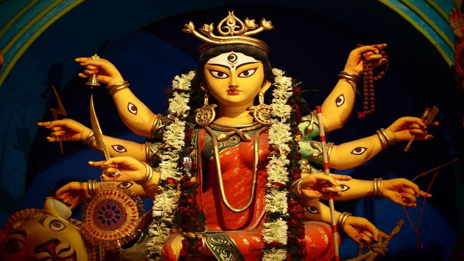 durga mata HD 桌布 壁紙 — 一尊色彩鮮豔、多臂的印度教女神雕像，飾有白花、金飾和紅衣，在戲劇性的藍色燈光下。