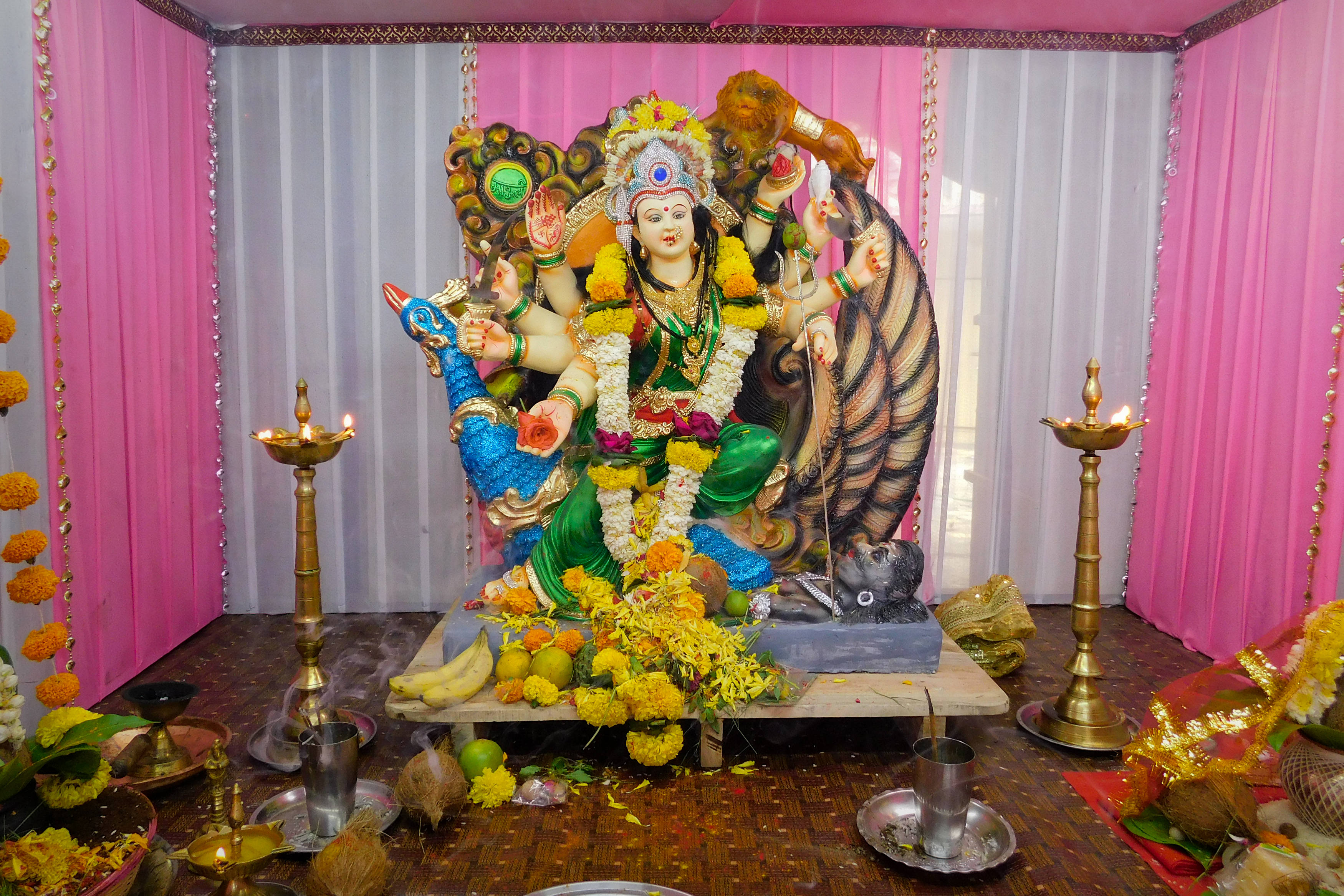 maa durga 桌布 壁紙 — 一尊色彩鮮豔、多臂的印度教女神雕像，飾有鮮花和供品，坐在粉紅色窗簾下裝飾華麗的祭壇上。