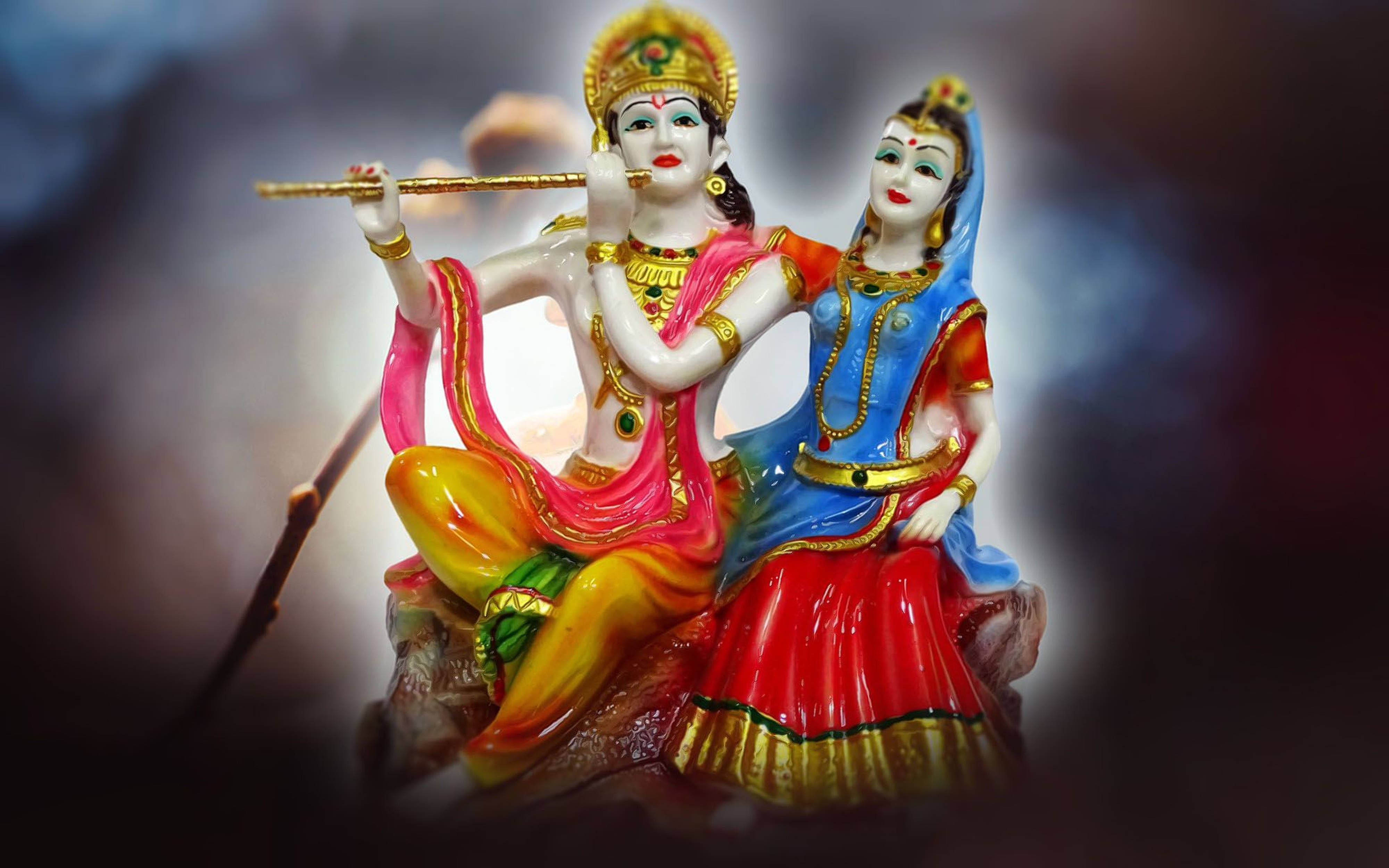 radha krishna 3d 桌布 壁紙 — 一尊色彩鮮豔的克里希納雕像在拉達旁邊吹奏長笛，身穿色彩繽紛的傳統服飾，背景柔和模糊。