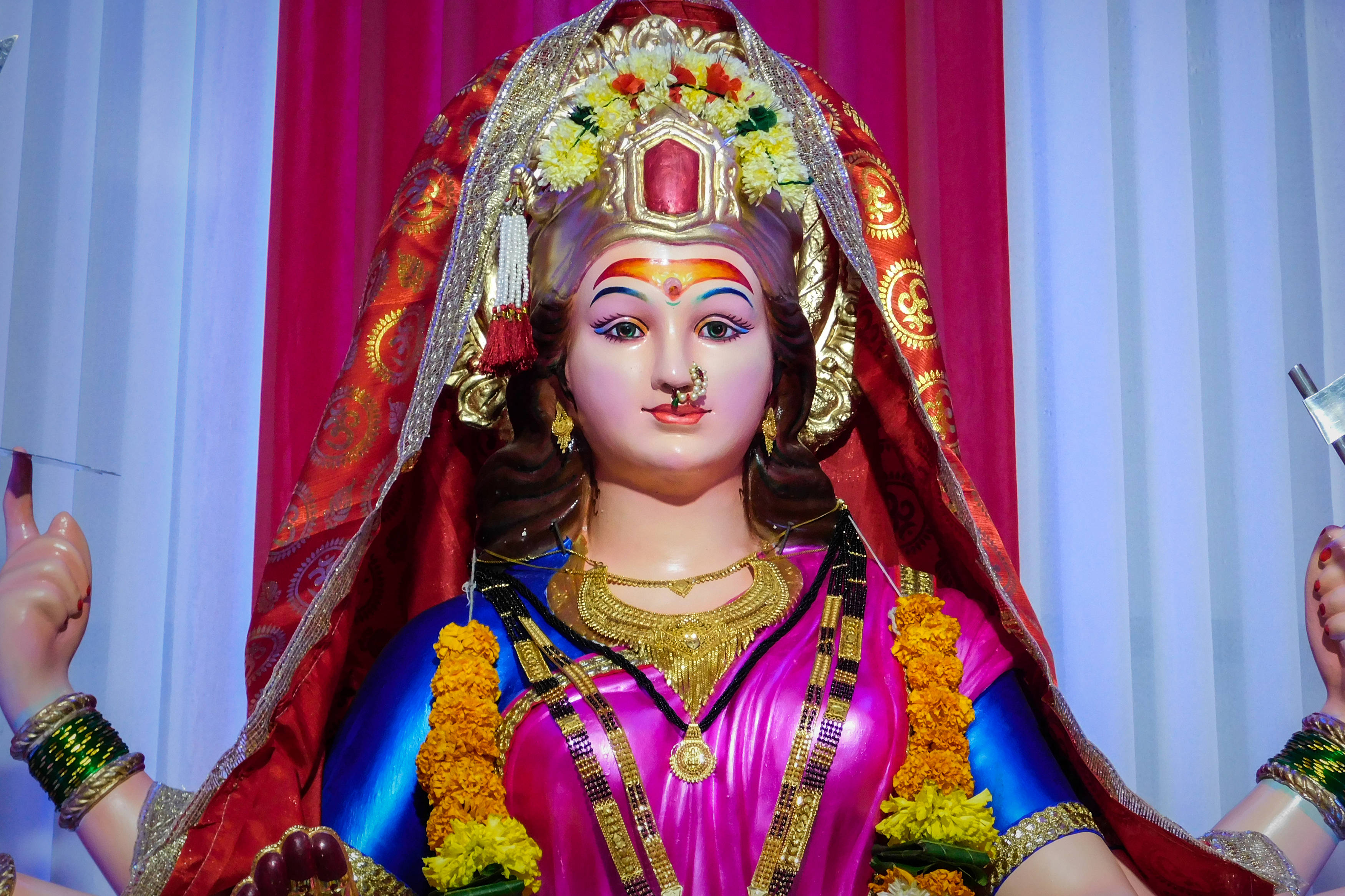 maa durga 桌布 壁紙 — 一尊色彩鮮豔的印度教女神雕像，以粉紅、藍色和金色裝飾，佩戴金盞花花環和精緻珠寶，背景為紅色和白色窗簾。