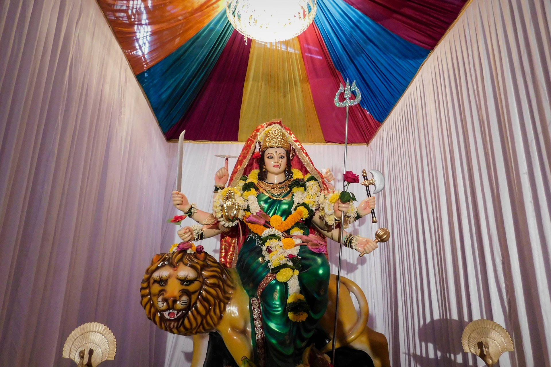 Durga Devi 桌布 壁紙 — 一尊色彩鮮豔的多臂女神雕像，身穿綠色服飾，騎著獅子，在色彩繽紛的垂簾天篷下，飾有鮮花和三叉戟。