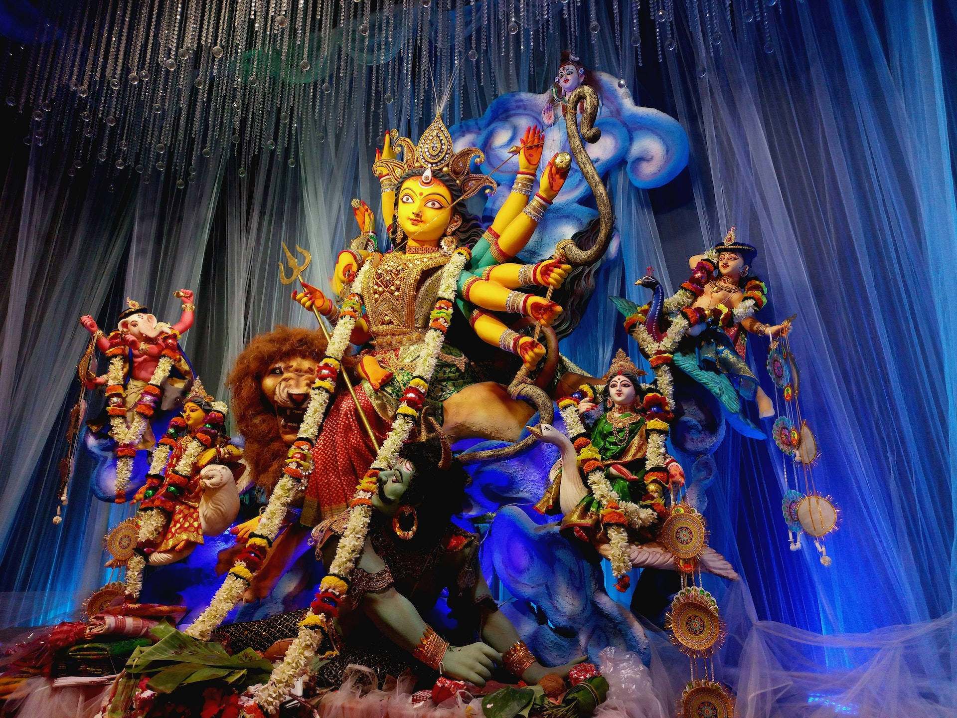 Durga Devi 桌布 壁紙 — 一尊色彩鮮豔的多臂女神雕像，飾有花環和武器，坐在獅子和孔雀上，周圍有侍從，背景是藍色垂簾。