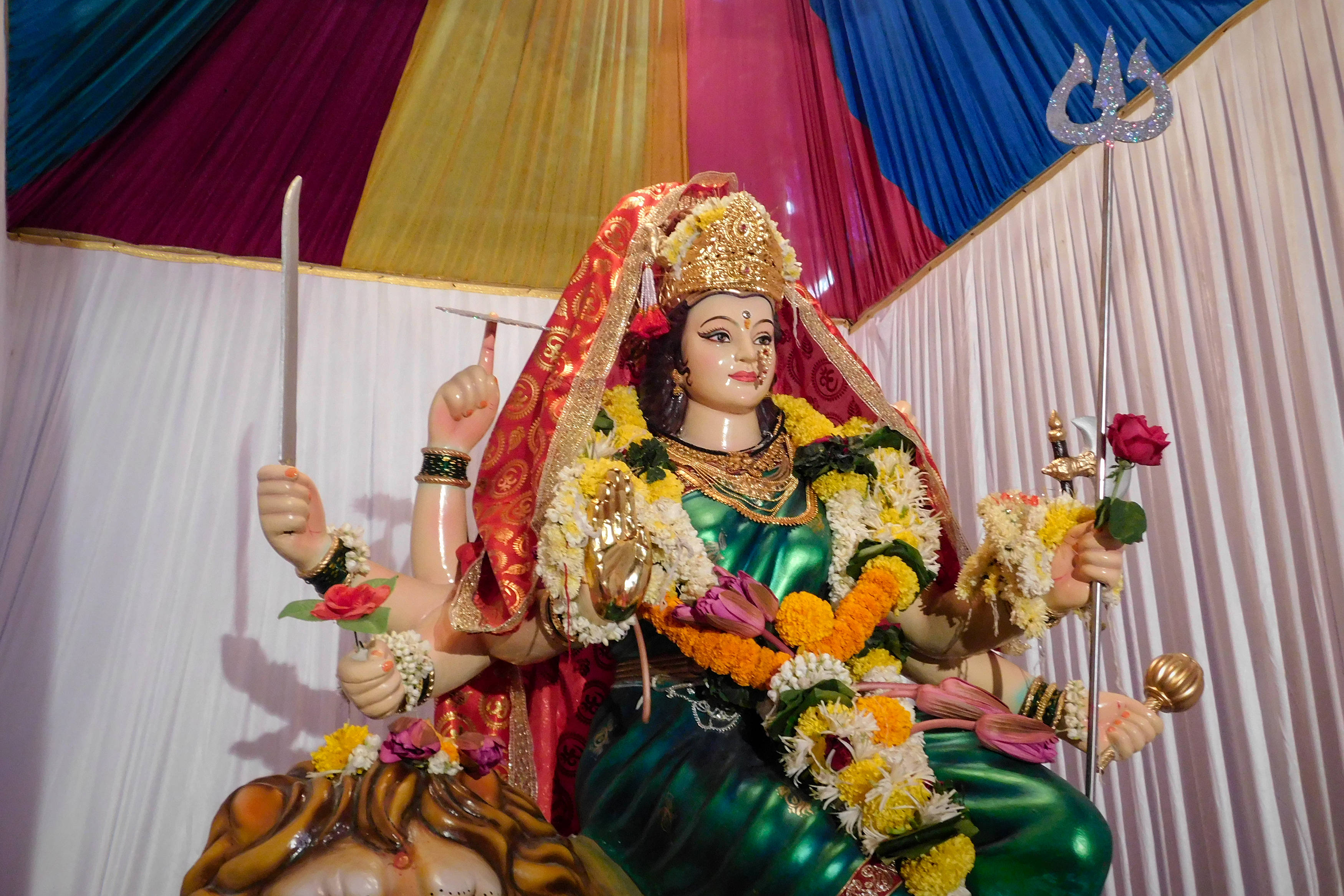Navratri 桌布 壁紙 — 一尊色彩鮮豔的杜爾迦女神雕像，飾有彩色花卉和珠寶，在多彩的布幔背景下手持武器。