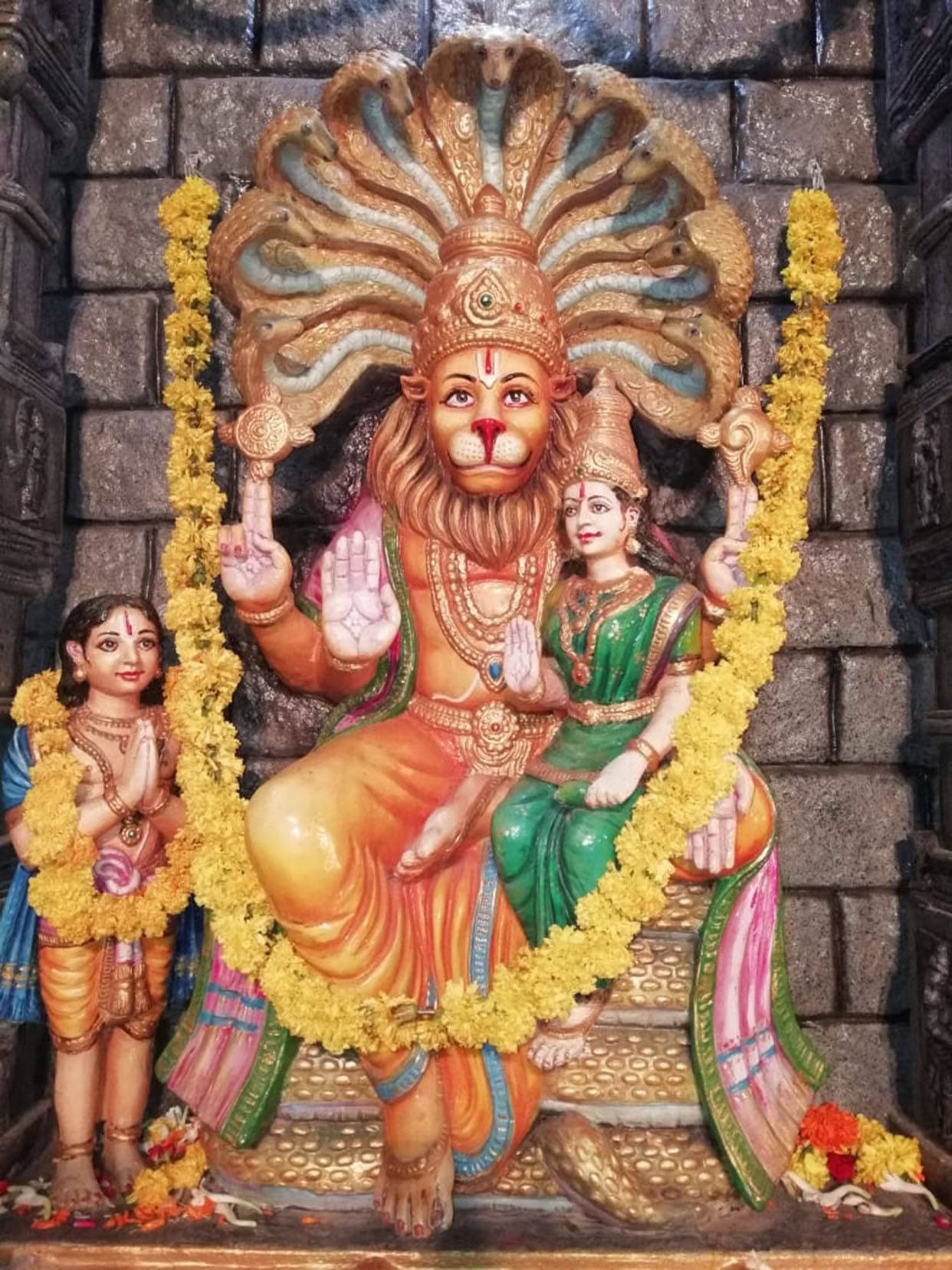 India 桌布 壁紙 — 一尊色彩鮮豔的納拉辛哈神像，獅頭人身，飾有金色和黃色花環，與配偶和孩子坐在寺廟環境中。
