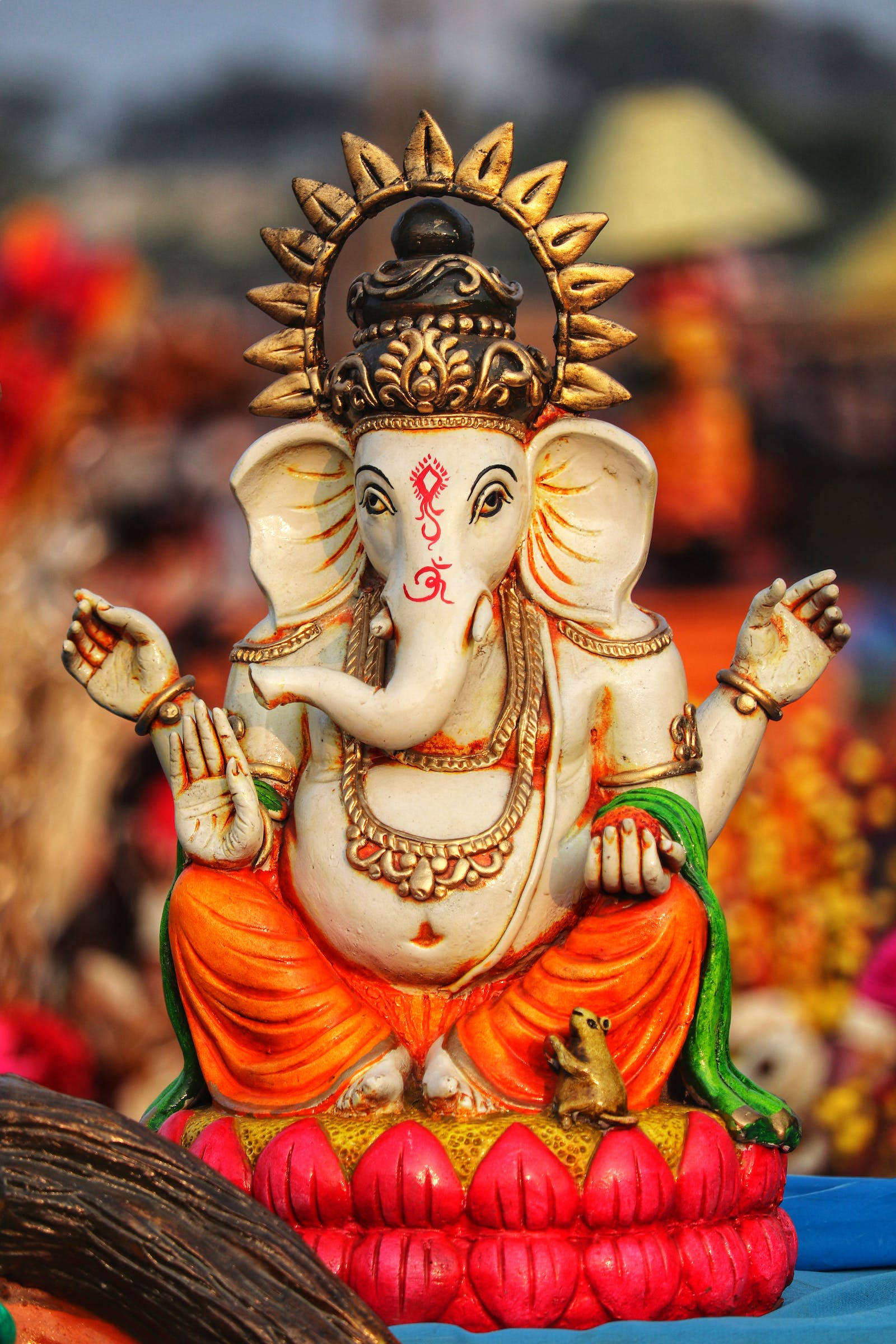 Ganesh 桌布 壁紙 — 一尊色彩鮮豔的象神雕像，飾有黃金首飾和橙色長袍，安詳地坐在粉紅色蓮花底座上。