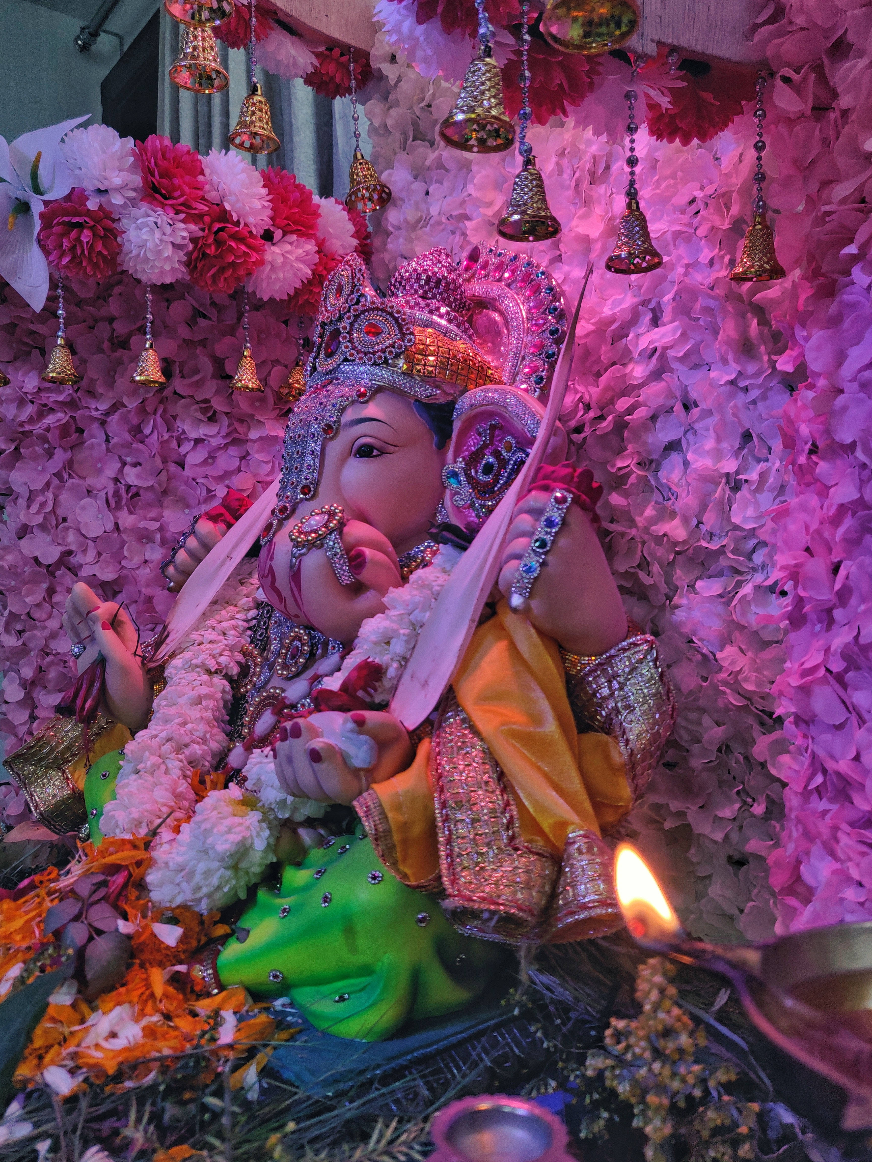 ganesh 4K 桌布 壁紙 — 一尊色彩鮮豔的象神雕像坐在粉紅色和白色花朵的瀑布中，附近有懸掛的鈴鐺和點燃的蠟燭。