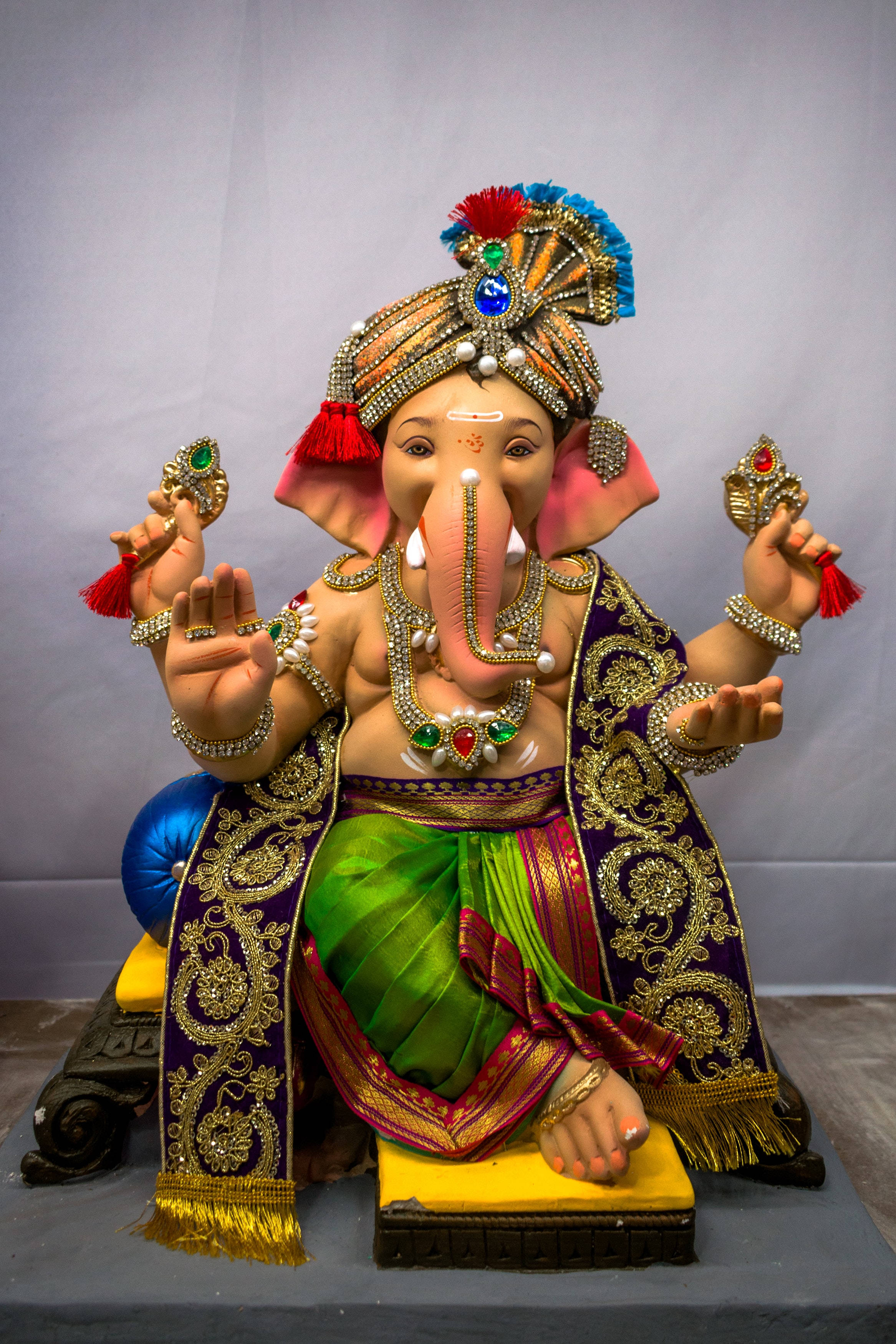 hindu iPhone 桌布 壁紙 — 一尊色彩鮮豔的象神雕像莊嚴地坐在寶座上，披著帶有金色刺繡和珠寶飾品的綠色和紫色布料。