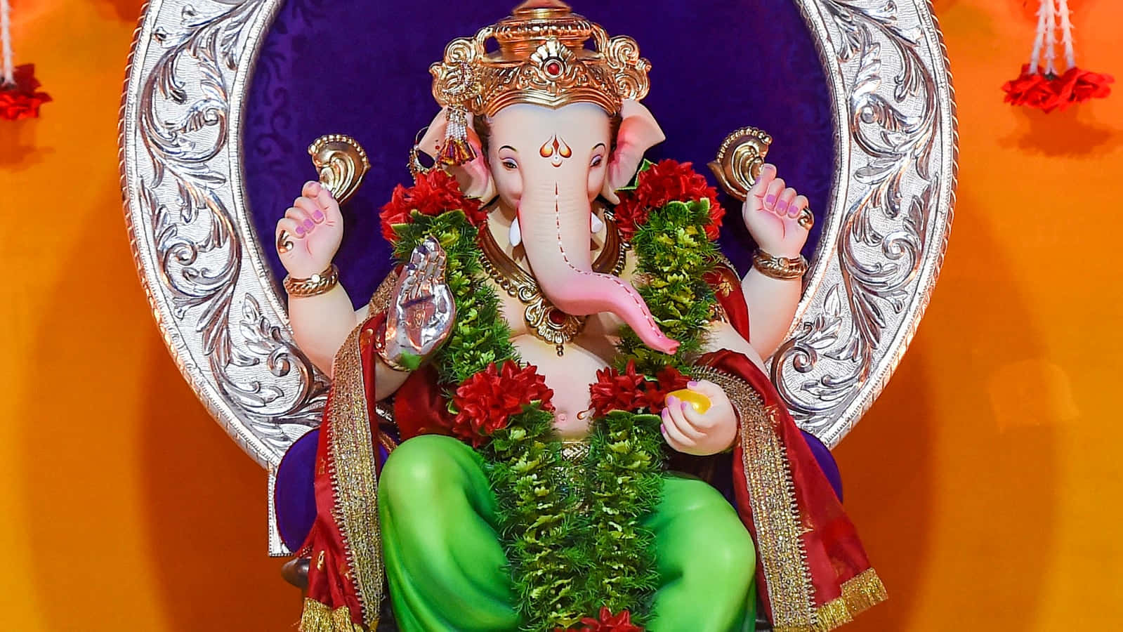 Ganesh 桌布 壁紙 — 一尊色彩鮮豔的象頭神像，飾有紅花和金飾，莊嚴地坐在紫色和橙色的背景前。