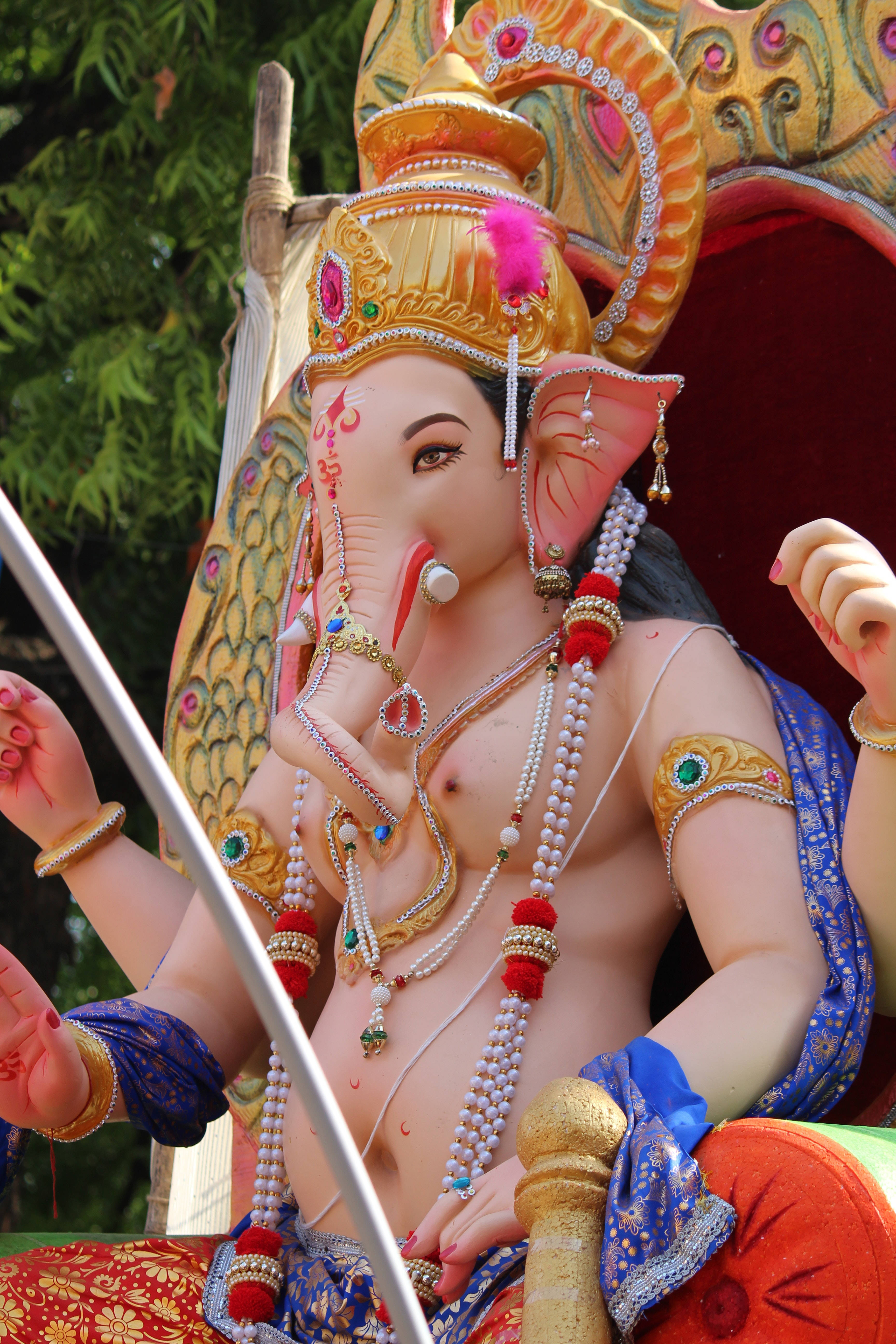 Ganesh 桌布 壁紙 — 一尊色彩鮮豔的金色象神雕像，飾有珠寶和藍色布料，端坐在綠色樹葉前，顯得莊嚴華麗。