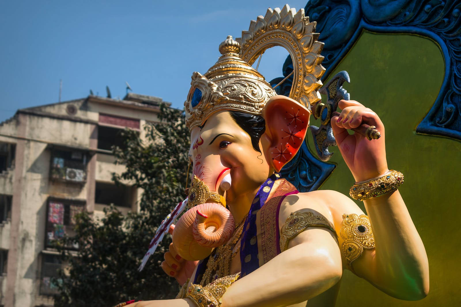 Ganesh 桌布 壁紙 — 一尊色彩鮮豔的金色象頭神像，擁有象頭和華麗王冠，在晴朗藍天下以城市建築為背景矗立。