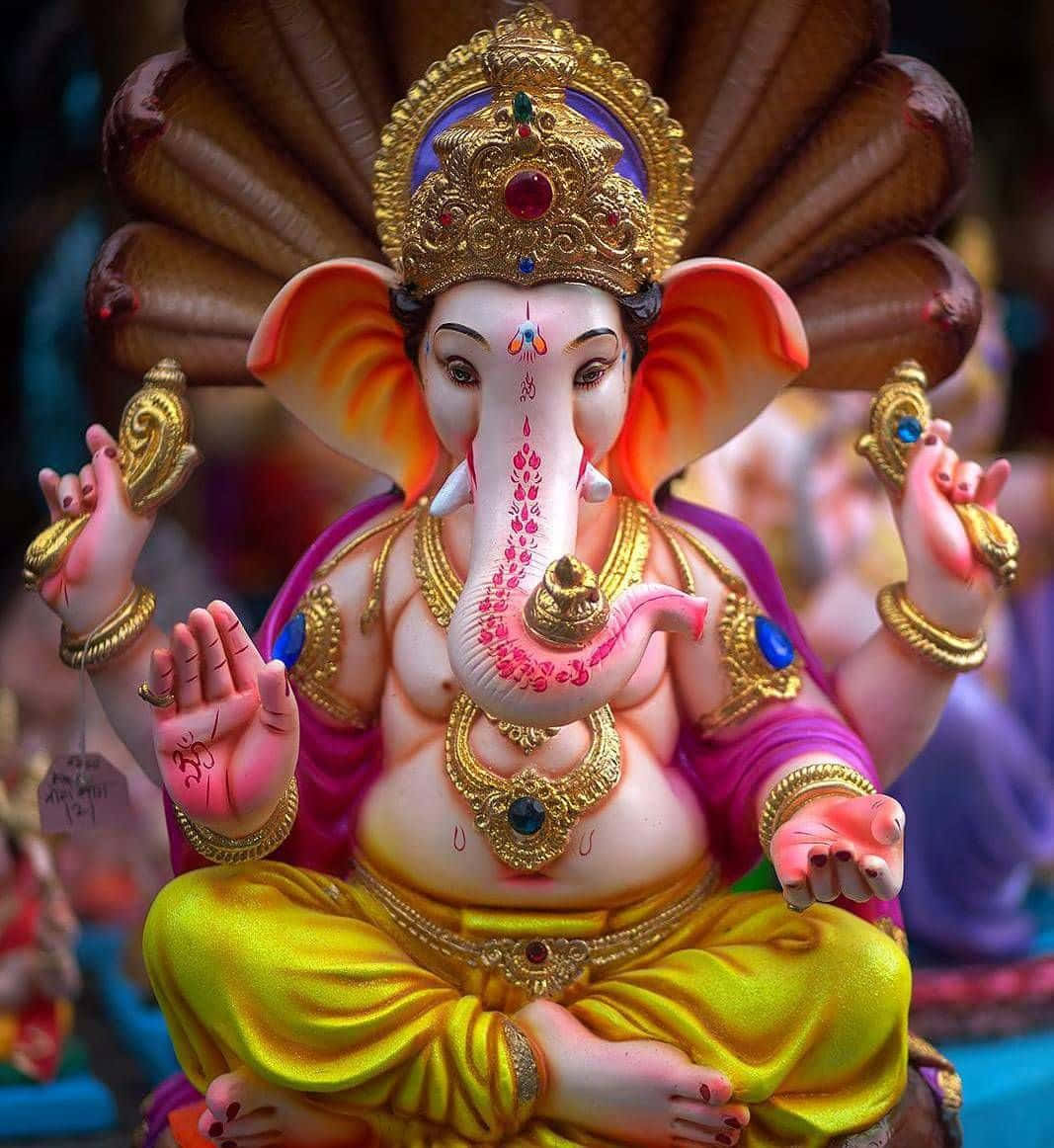 Ganesh 桌布 壁紙 — 一尊色彩鮮豔的金色象頭神像，粉紅色皮膚，佩戴華麗珠寶，以冥想姿勢坐著。