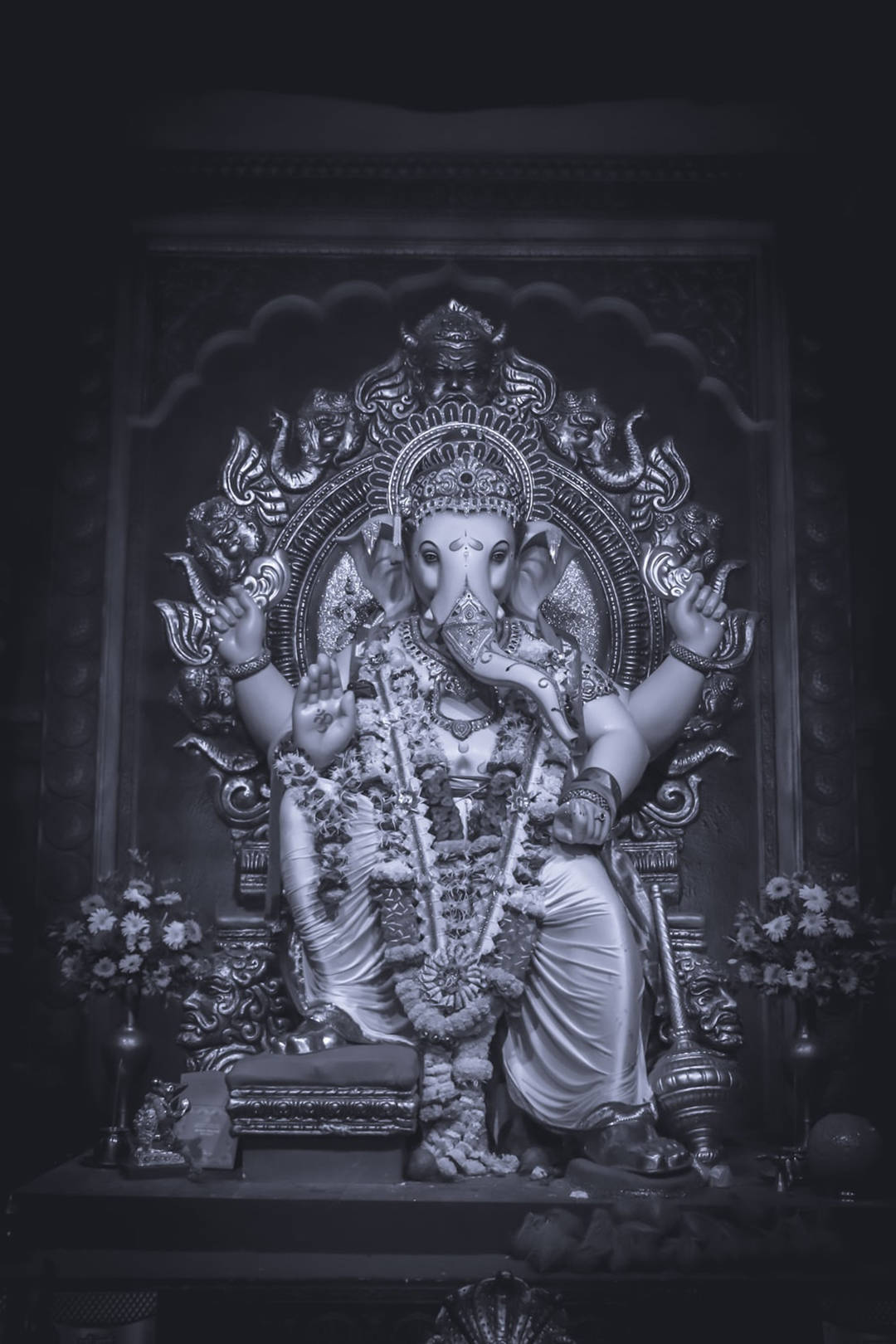 Ganesh 桌布 壁紙 — 一尊莊嚴的單色象頭神雕像，飾以花環，威嚴地坐在華麗的寶座上，周圍環繞著精緻的雕刻和花卉供品。