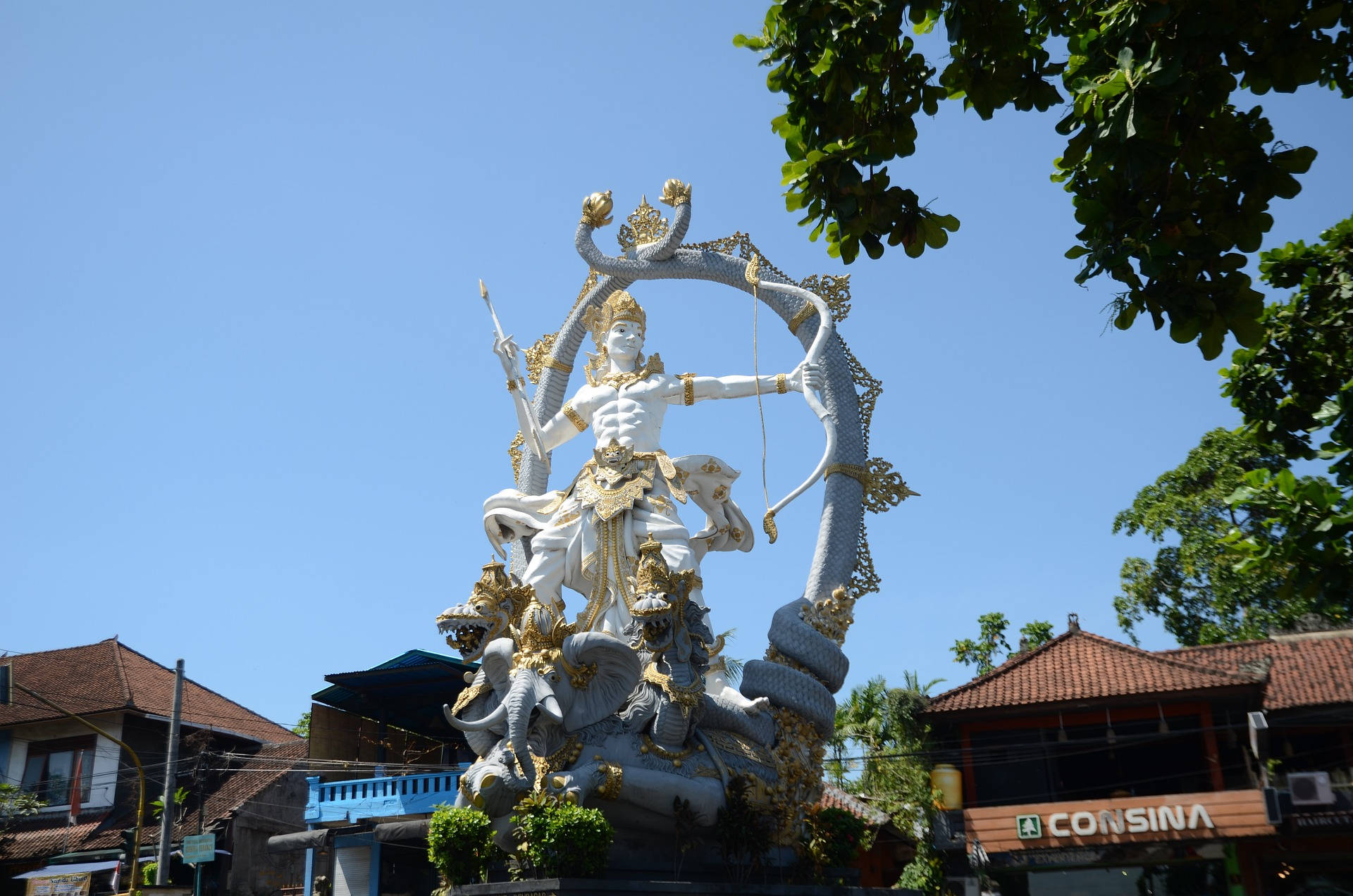 shiv shankar 桌布 壁紙 — 一尊莊嚴的白金色神像騎著多頭蛇，背景是清澈的藍天，茂密的綠樹框住了整個場景。