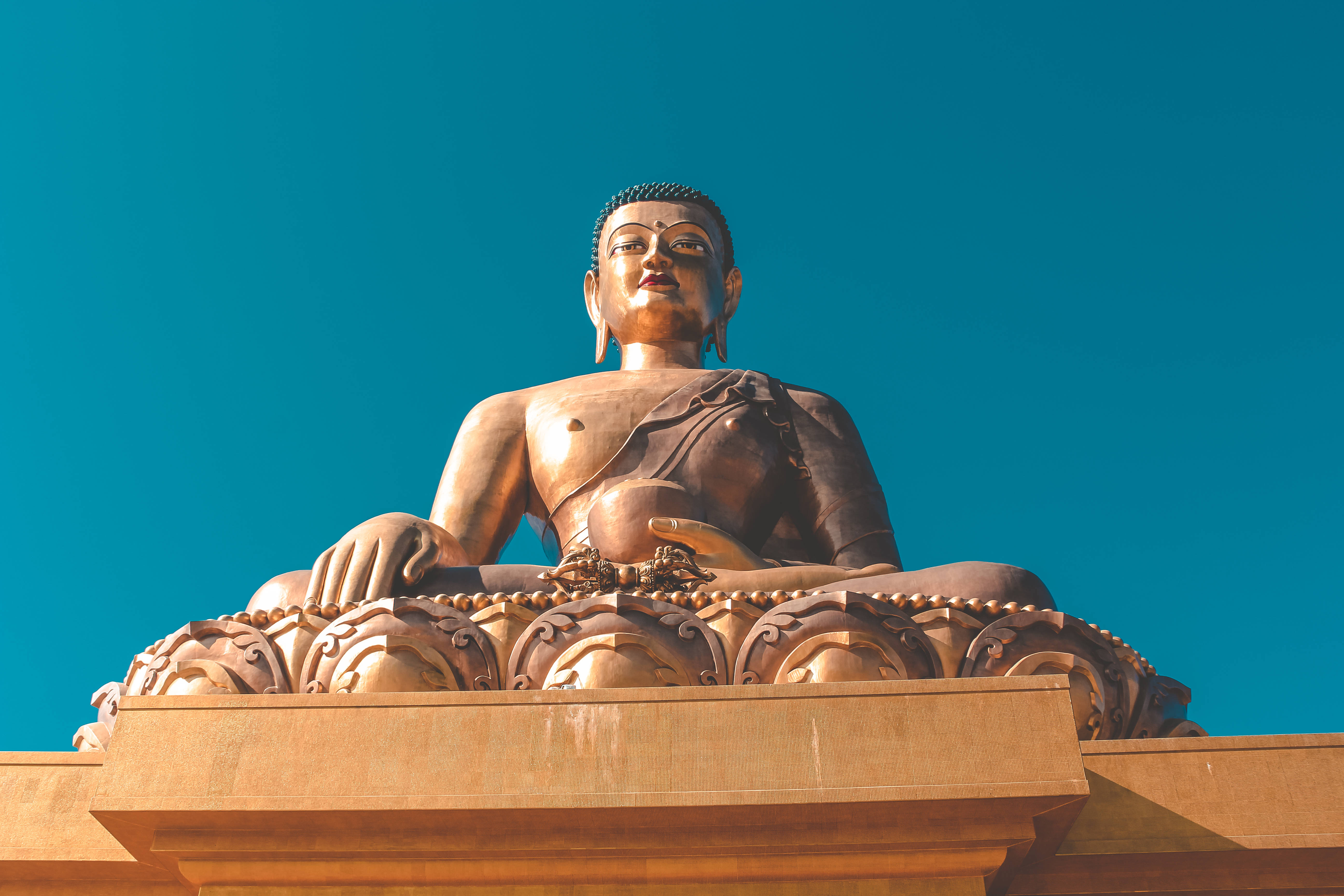 Bhutan 桌布 壁紙 — 一尊莊嚴的金色佛像，在鮮豔的藍綠色天空下，安詳地坐在華麗的基座上。