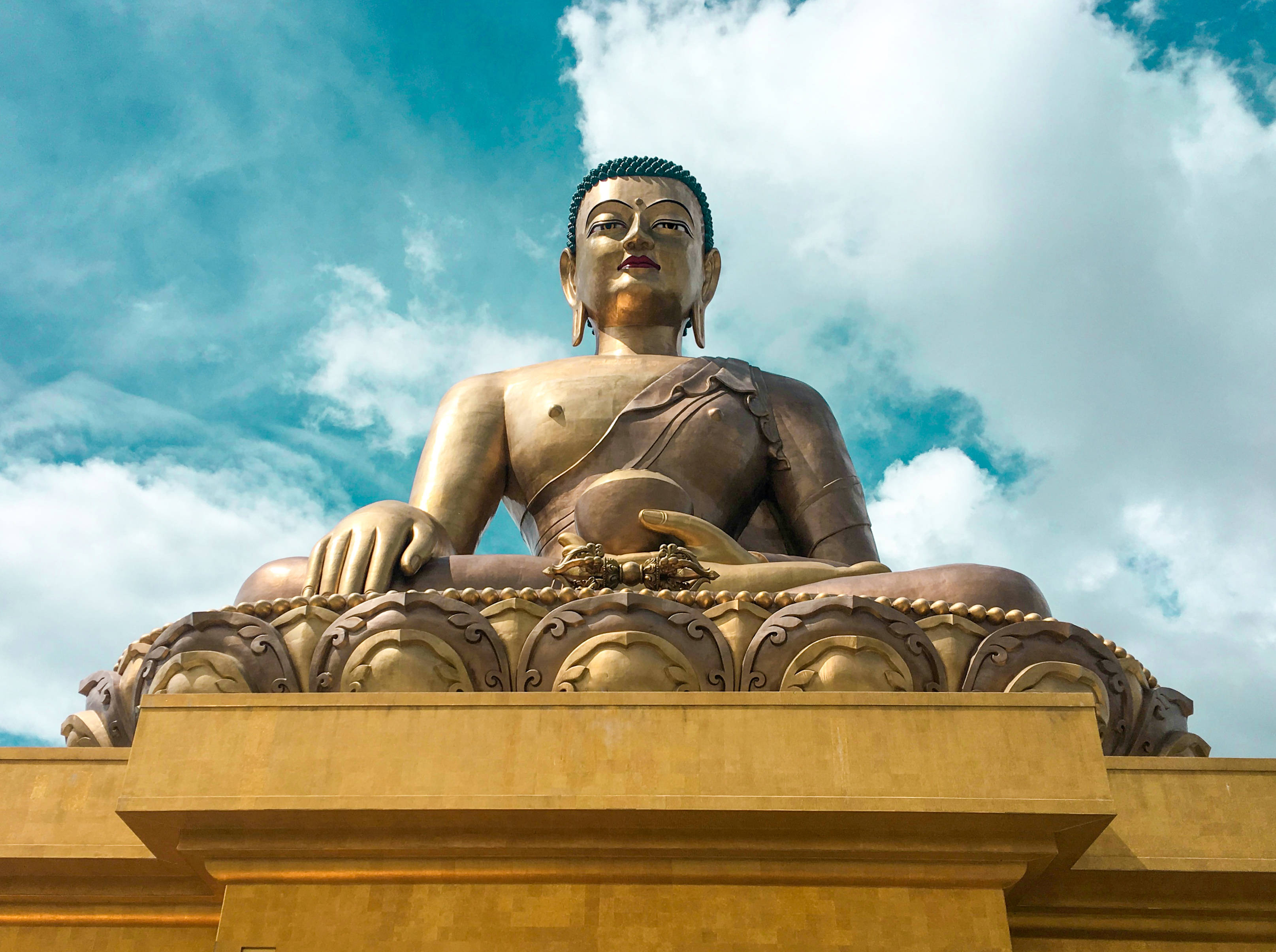 Bhutan 桌布 壁紙 — 一尊莊嚴的金色佛像安詳地坐在基座上，背景是充滿蓬鬆白雲的鮮豔藍天。