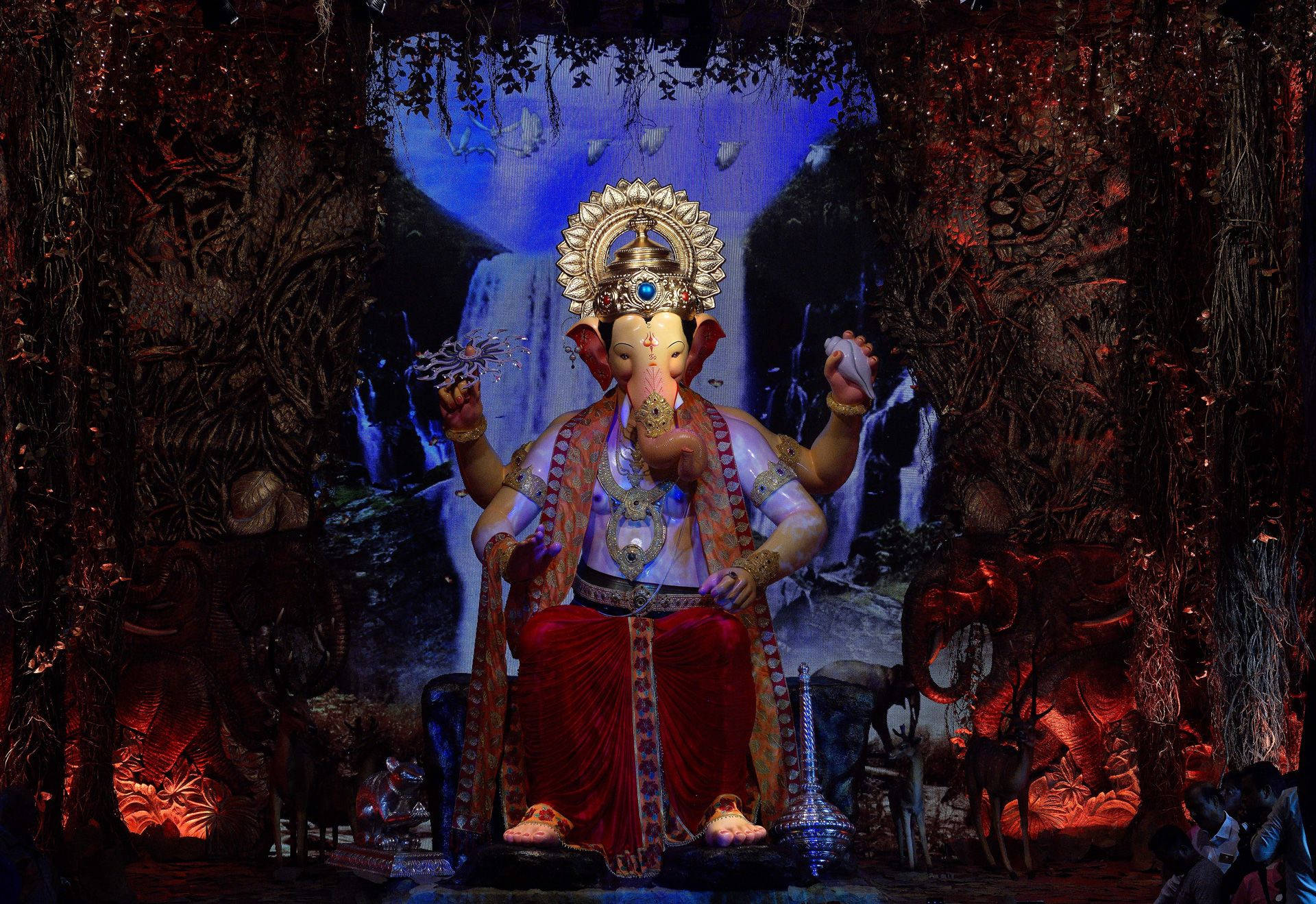 Hindu Temple 桌布 壁紙 — 一尊莊嚴的金色象神雕像，飾以紅色和金色，端坐在由深色樹葉環繞的發光瀑布背景前。