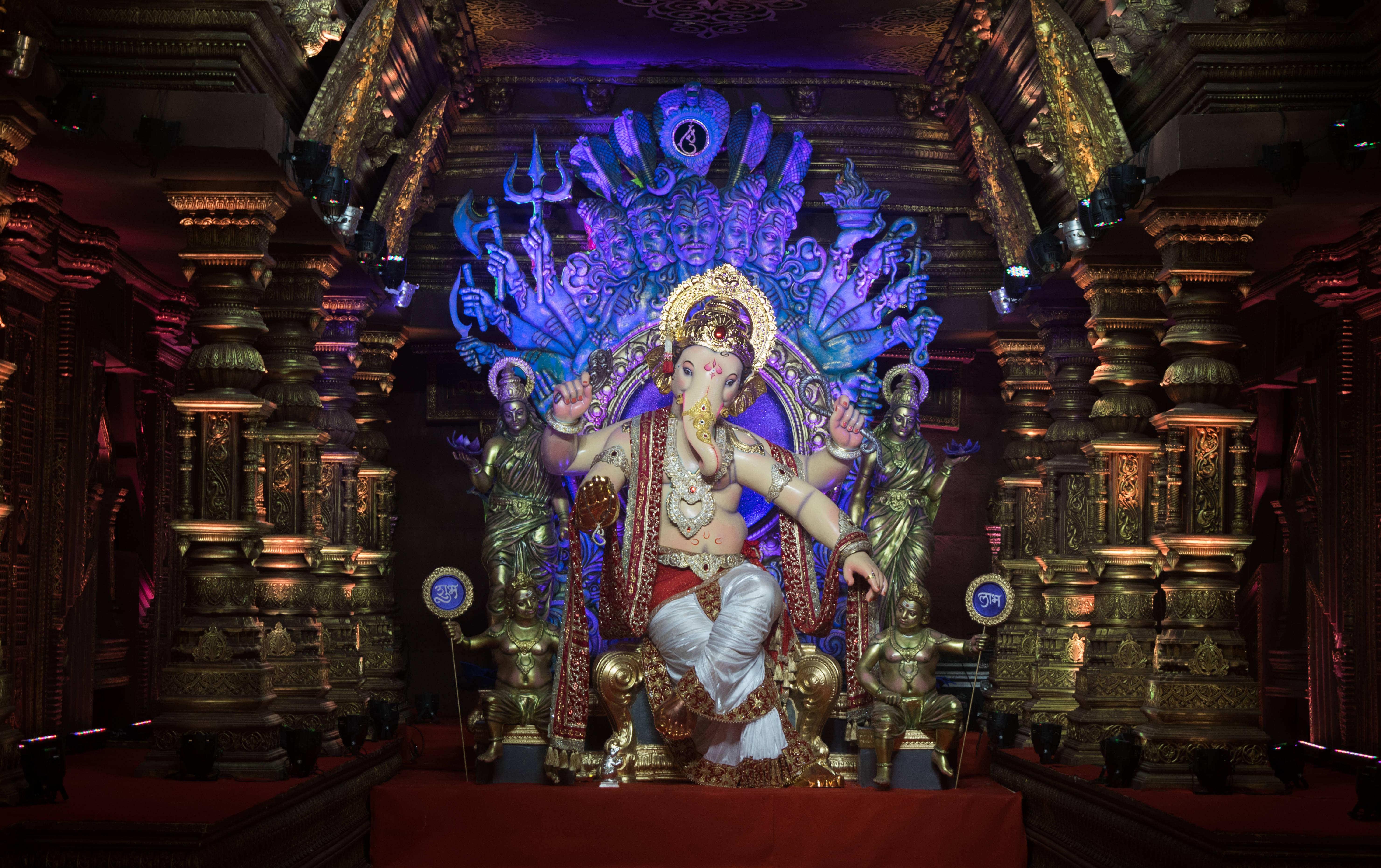 ganpati HD 桌布 壁紙 — 一尊莊嚴的金色象頭神雕像，有多隻手臂和耀眼的藍色光環，坐在裝飾華麗的寺廟寶座上，周圍有發光的紅色和金色柱子。
