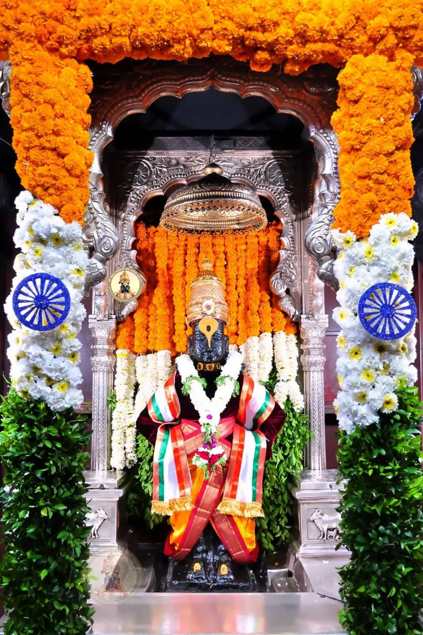 Hindu Temple 桌布 壁紙 — 一尊莊嚴的黑色神像，飾以橙色萬壽菊花環和印度三色肩帶，矗立在裝飾性的銀色拱門下，拱門由白色花朵和藍色阿育王法輪環繞。