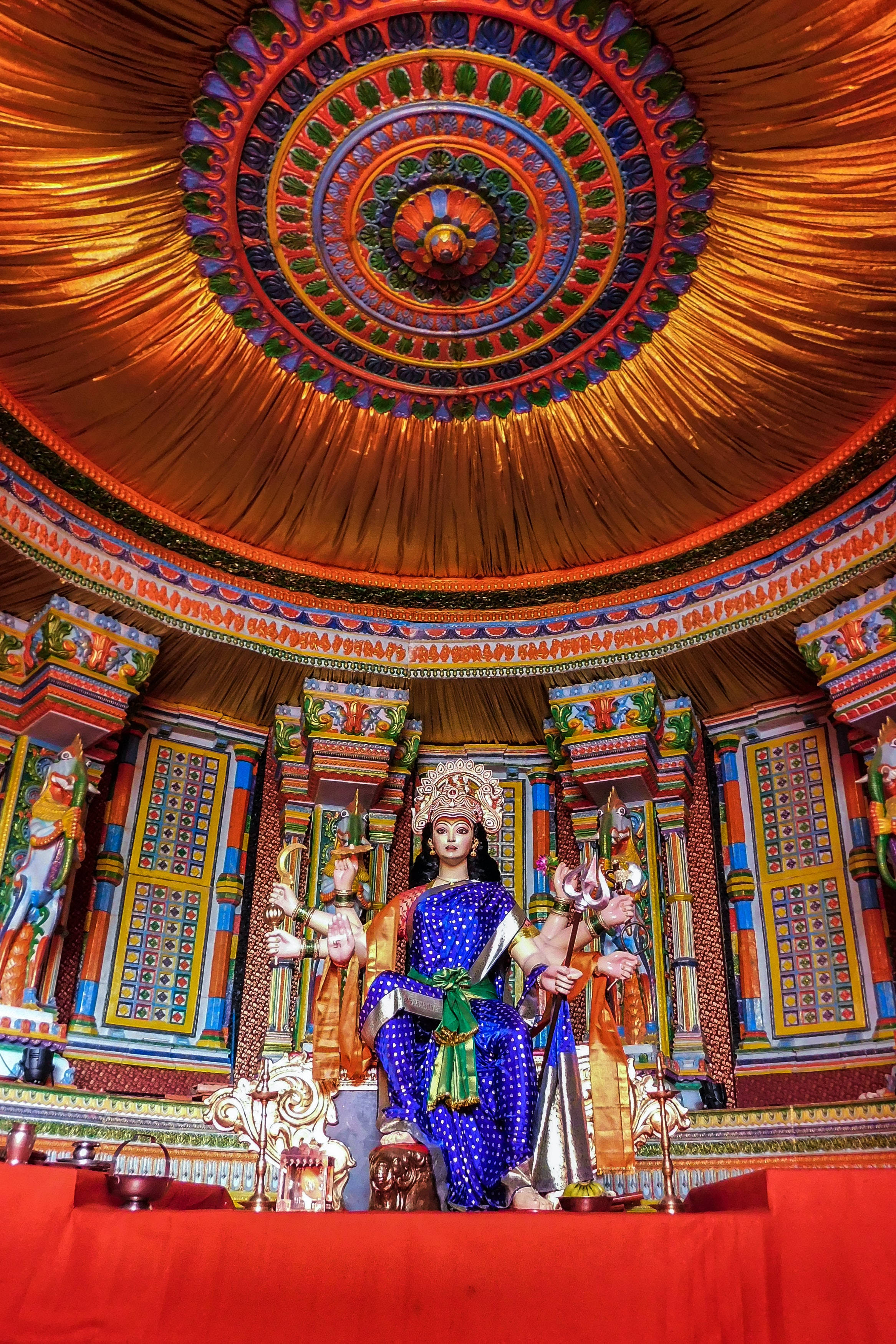 Mata Rani 桌布 壁紙 — 一尊華麗裝飾的藍金色神像，坐落在裝飾華麗的圓形天花板下，配有耀眼的橙色帷幔。