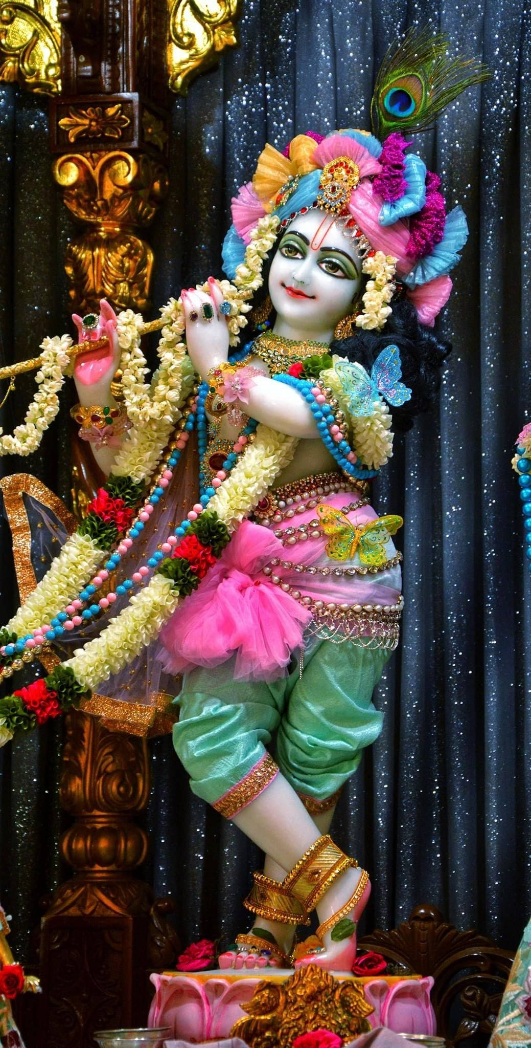 krishna bhagwan 桌布 壁紙 — 一尊裝飾華麗的克里希納神像，在閃爍的深色窗簾背景下吹奏長笛，飾有色彩繽紛的花朵、孔雀羽毛和金飾。