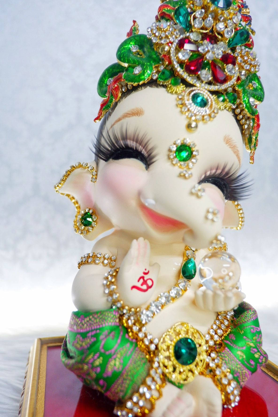 cute ganesha 桌布 壁紙 — 一尊裝飾華麗的嬰兒象神雕像，戴著鑲有寶石的皇冠，身穿綠色金色服飾，在柔和的白色背景前溫暖地微笑。