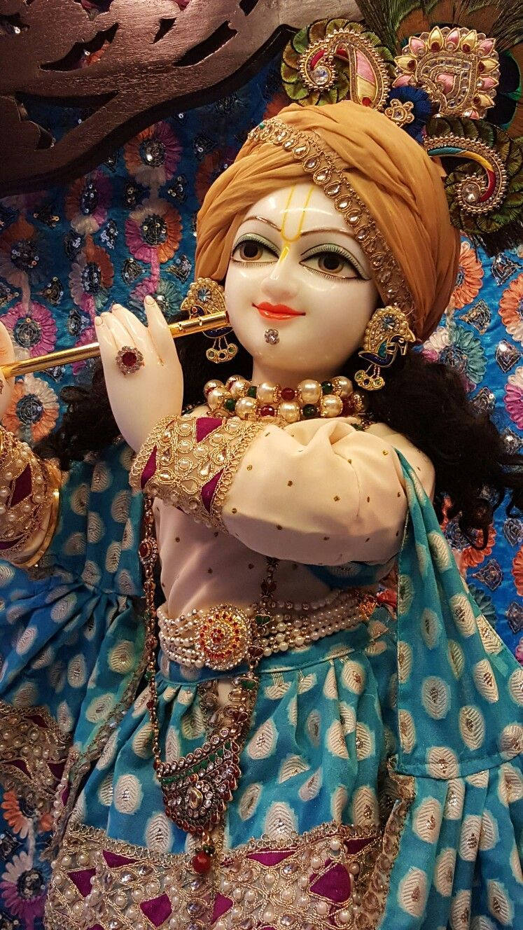 krishna god 桌布 壁紙 — 一尊裝飾華麗的神像，戴著金色頭巾，身穿藍色圖案服飾，在花卉背景前吹奏長笛。