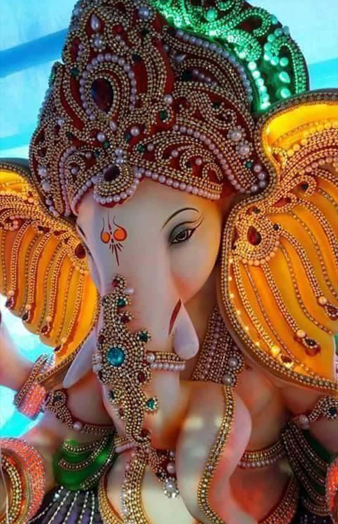 Ganesh 桌布 壁紙 — 一尊裝飾華麗的象神雕像，飾有金色珠寶、綠色珍珠和精緻的珠飾工藝，背景為柔和的藍色。