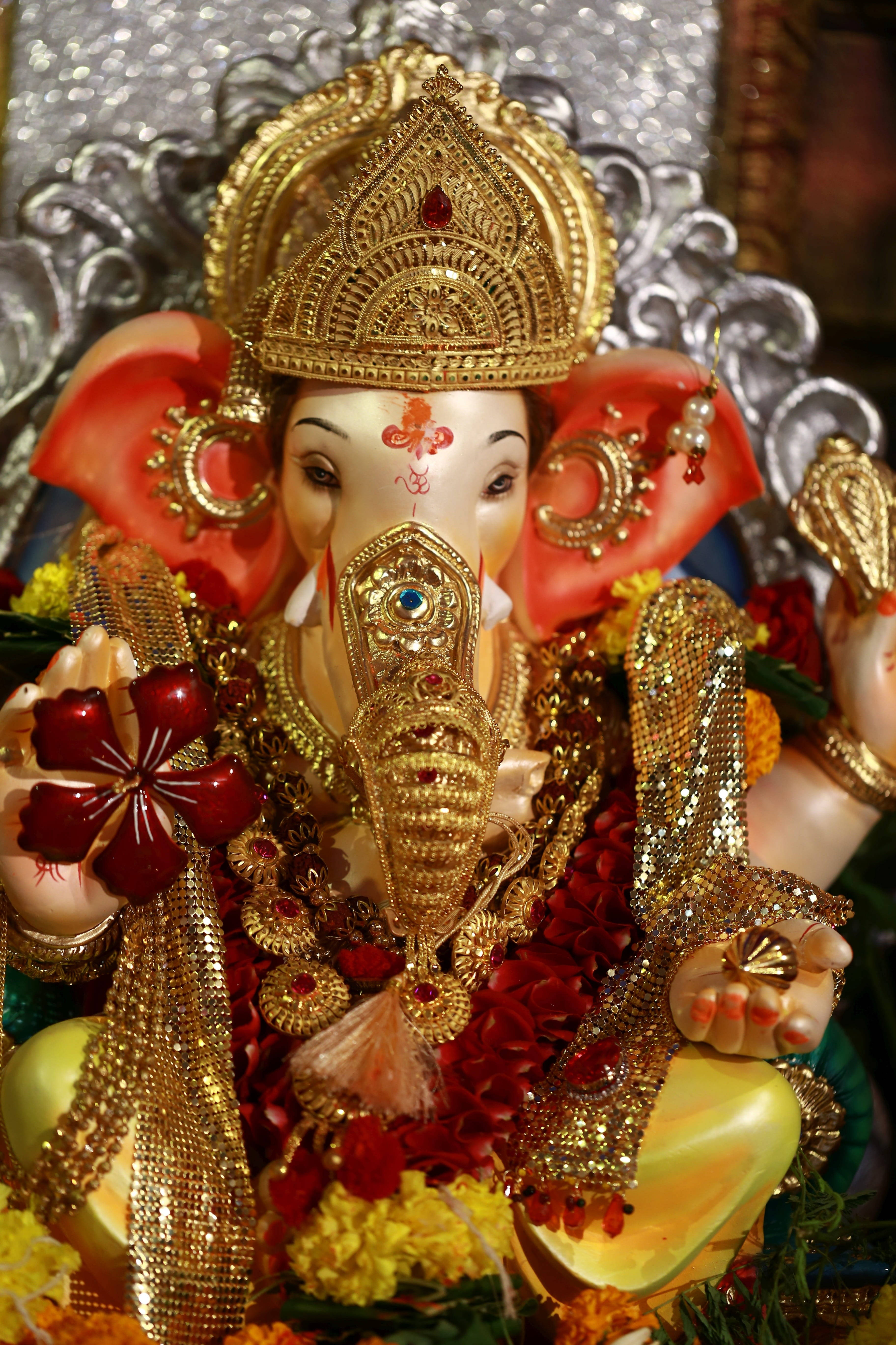 ganesh iPhone 桌布 壁紙 — 一尊裝飾華麗的黃金象頭神像，紅耳戴花環，莊嚴地坐在閃爍的銀色背景前。